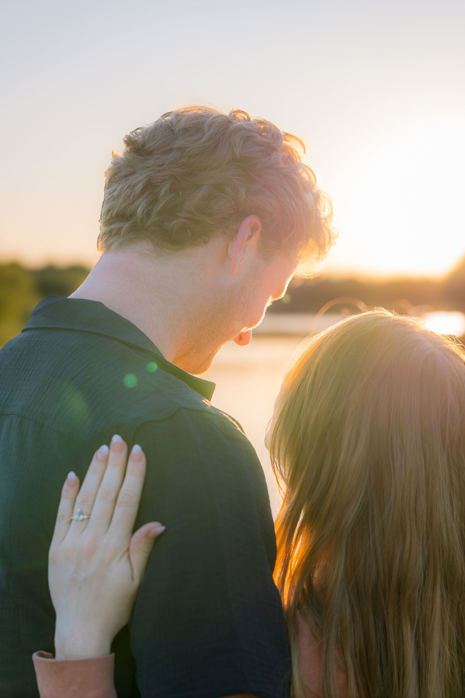 Couple embracing at golden sunset by the water, woman with ring resting hand on mans shoulder.