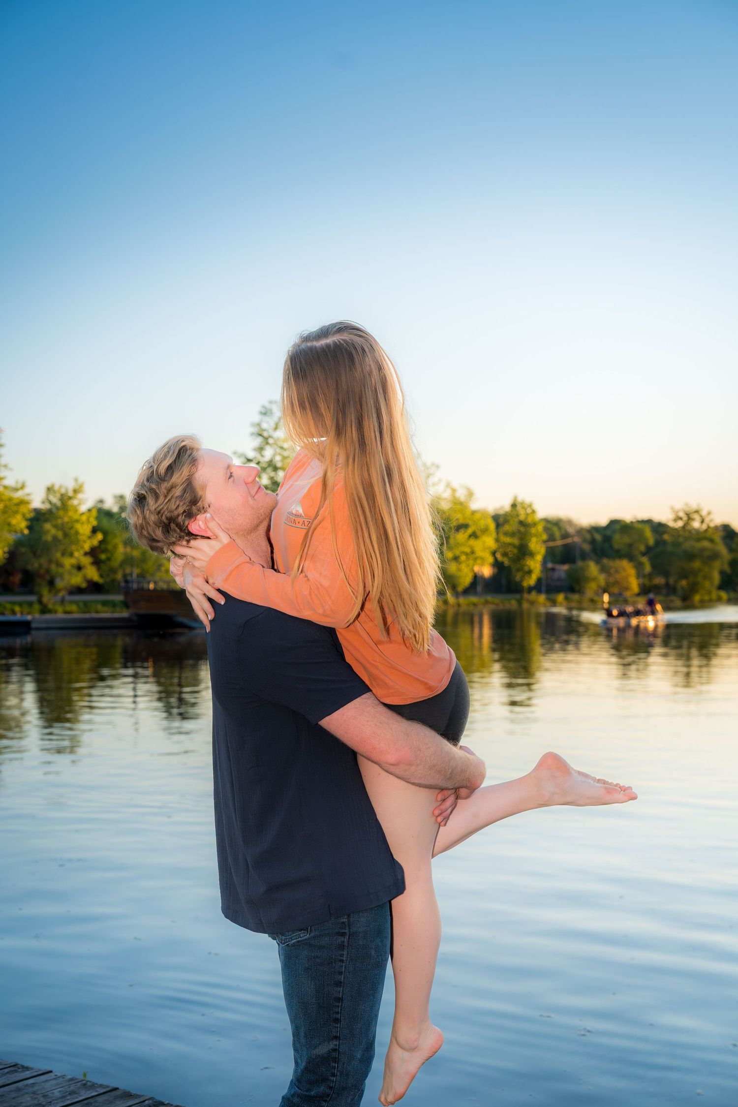 Man lifting and kissing woman on a dock by a peaceful lake at sunset.