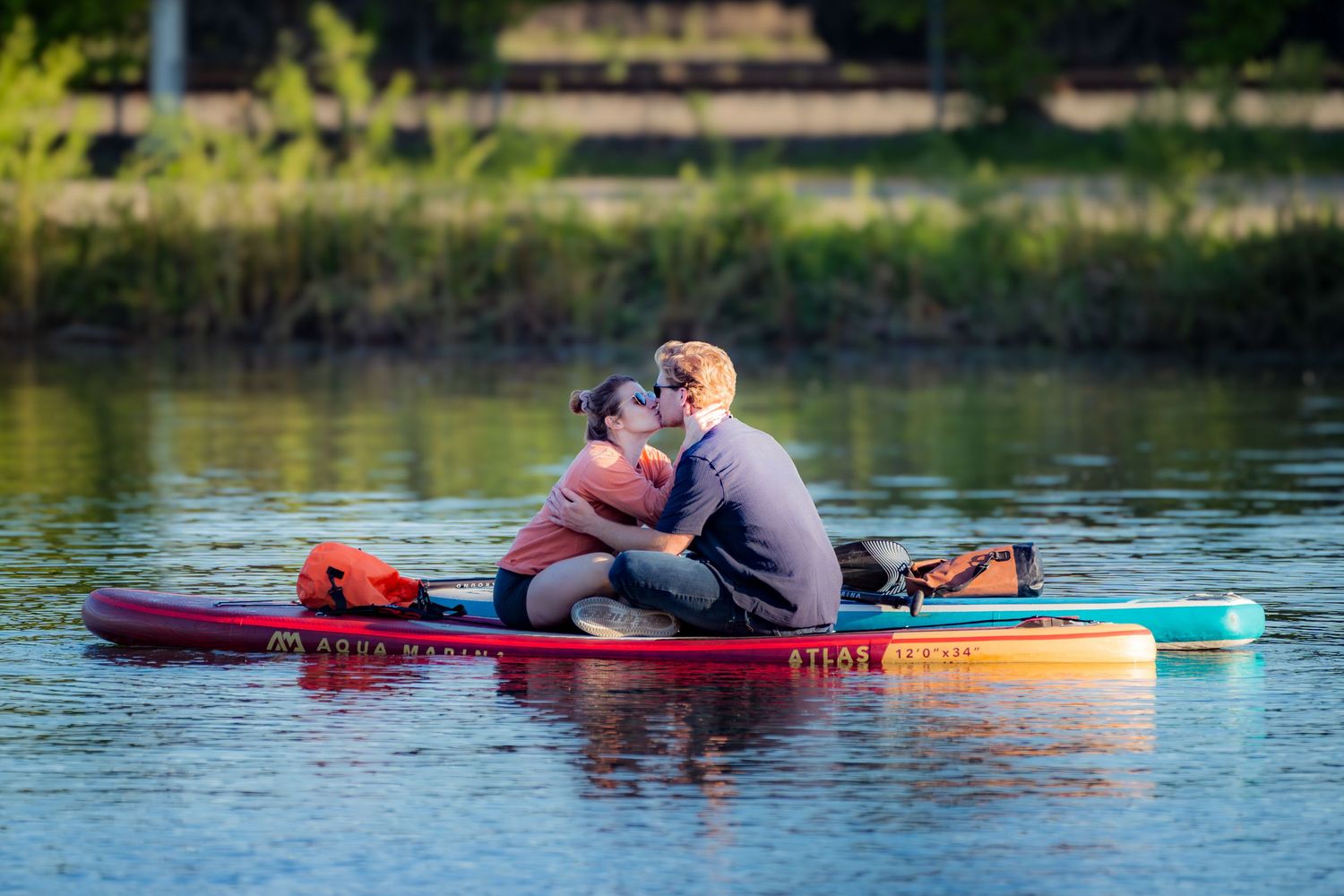Couple sharing a kiss while sitting together on a red paddleboard on a serene lake.