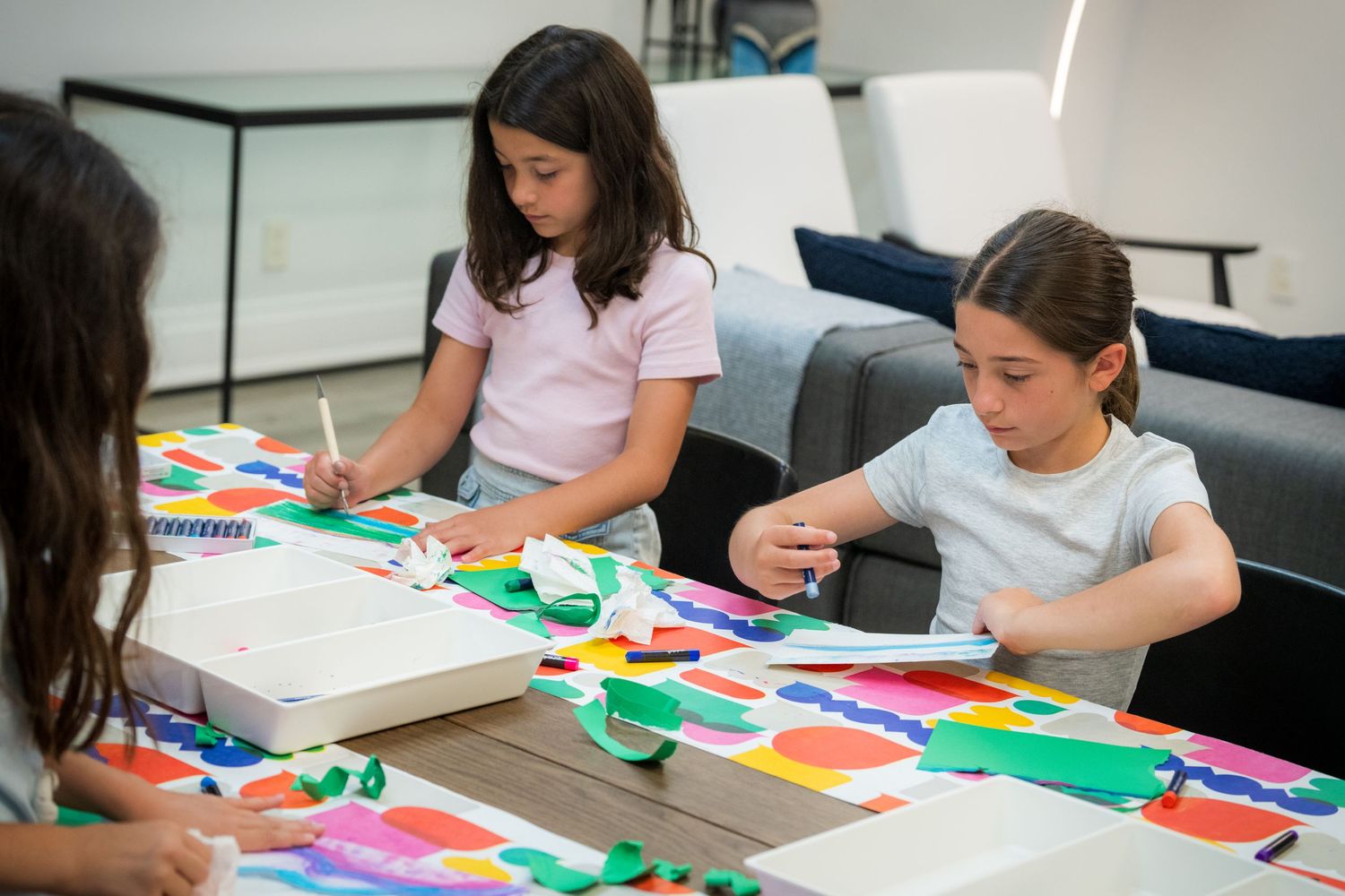 Three girls work together on colorful paper collage art project at table during creative workshop.