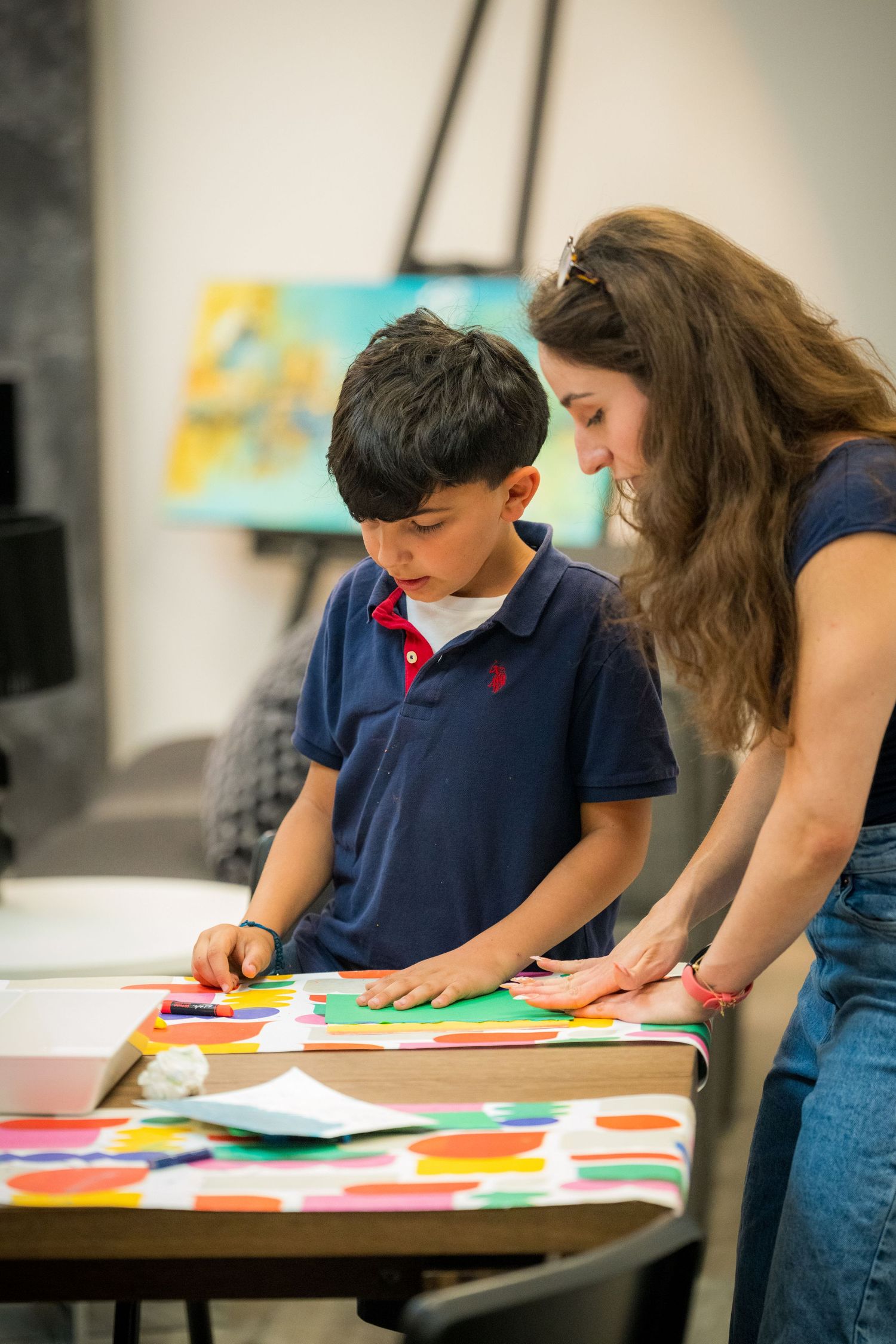 Woman assists young boy with colorful paper craft project at an art table.