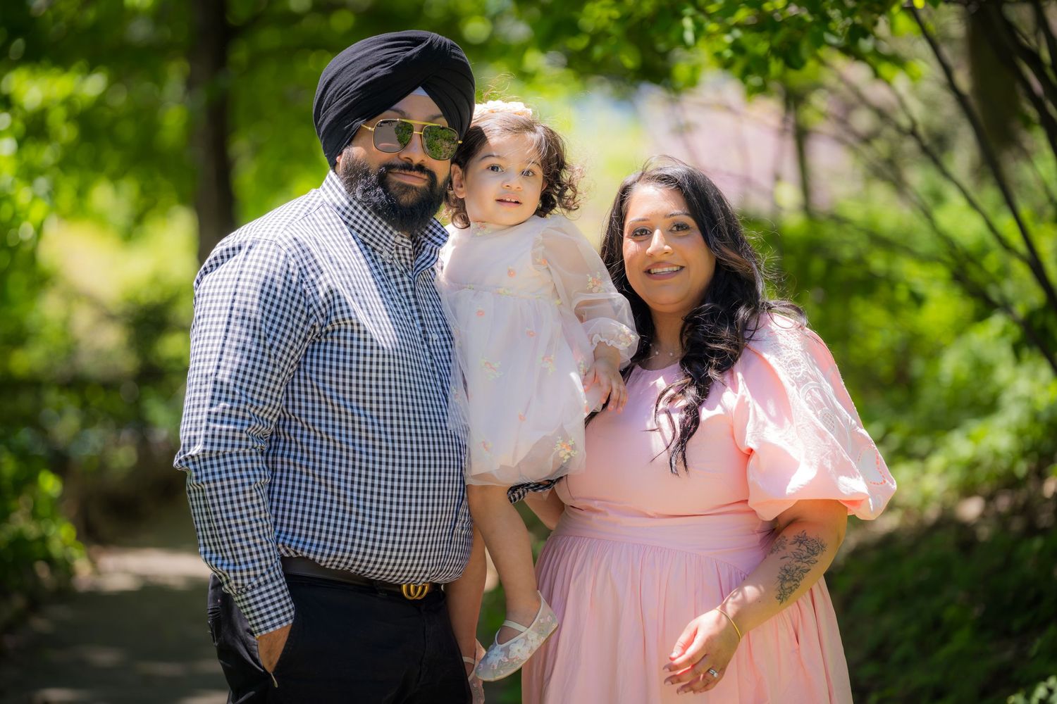 A Sikh family with a man in turban, woman in pink dress, and toddler posing in a green park.
