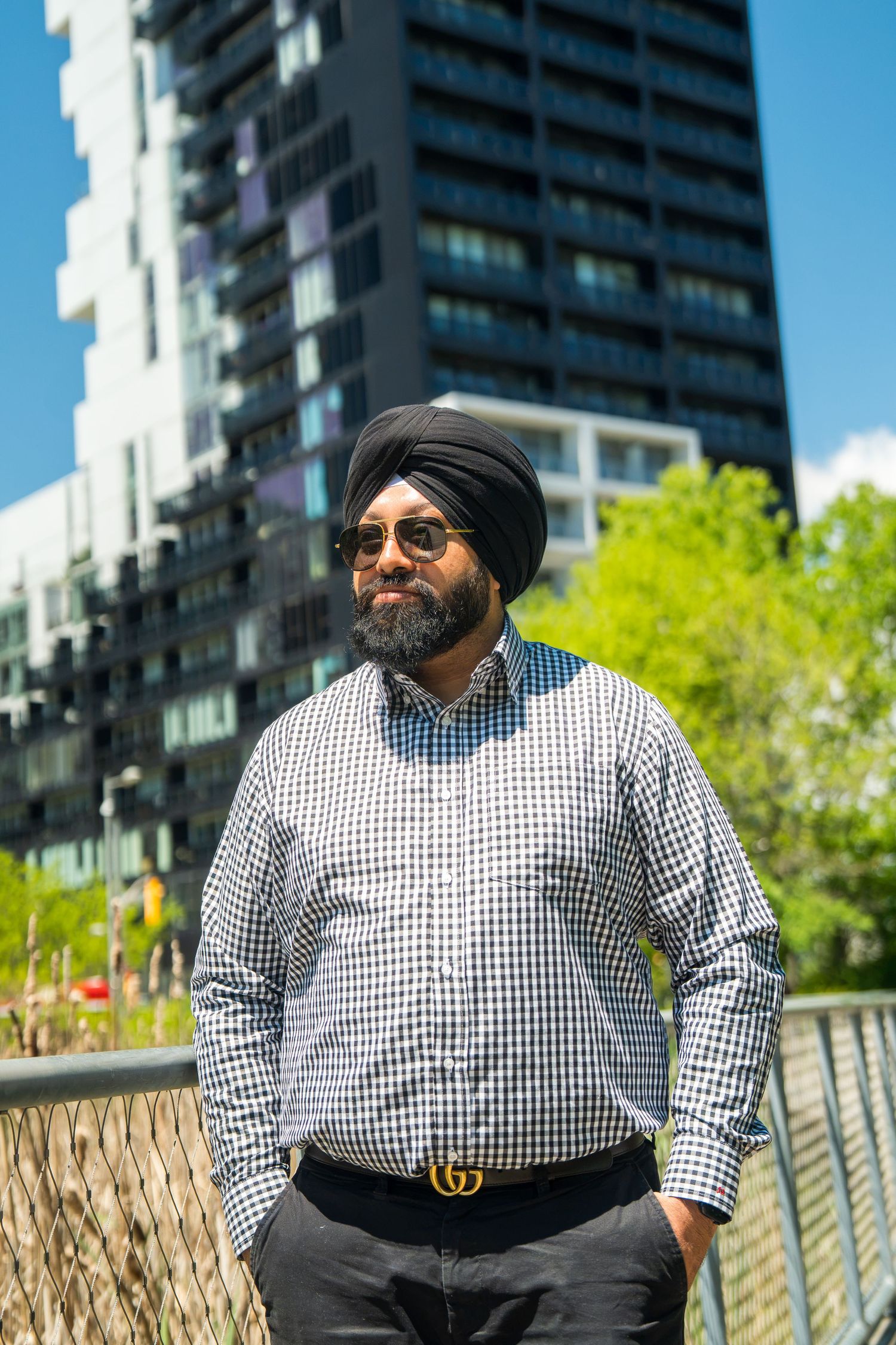 A bearded Sikh man wearing a black turban and checkered shirt stands confidently in front of a modern building.