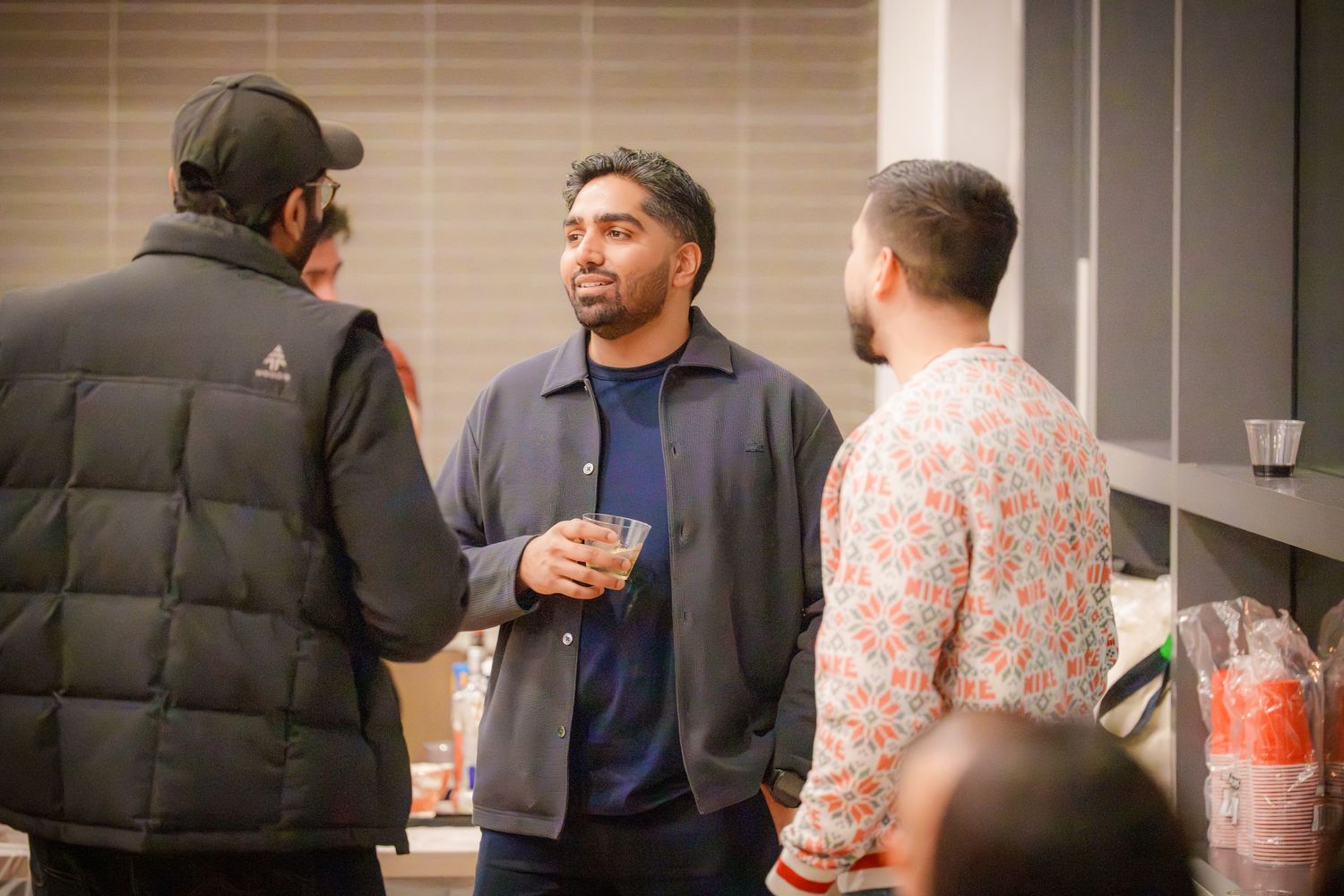 People mingling and socializing at an indoor party with window blinds in the background.
