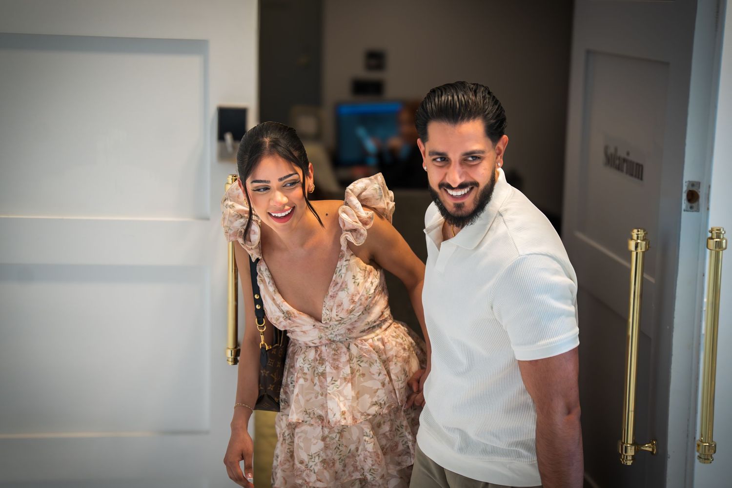 Smiling couple holding hands posing in a doorway, woman in floral dress and man in white shirt.