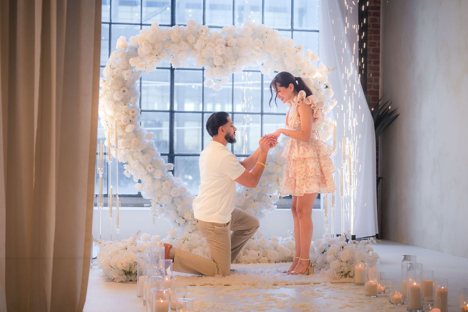 Man kneeling to propose to his girlfriend under a glowing heart-shaped floral arch.