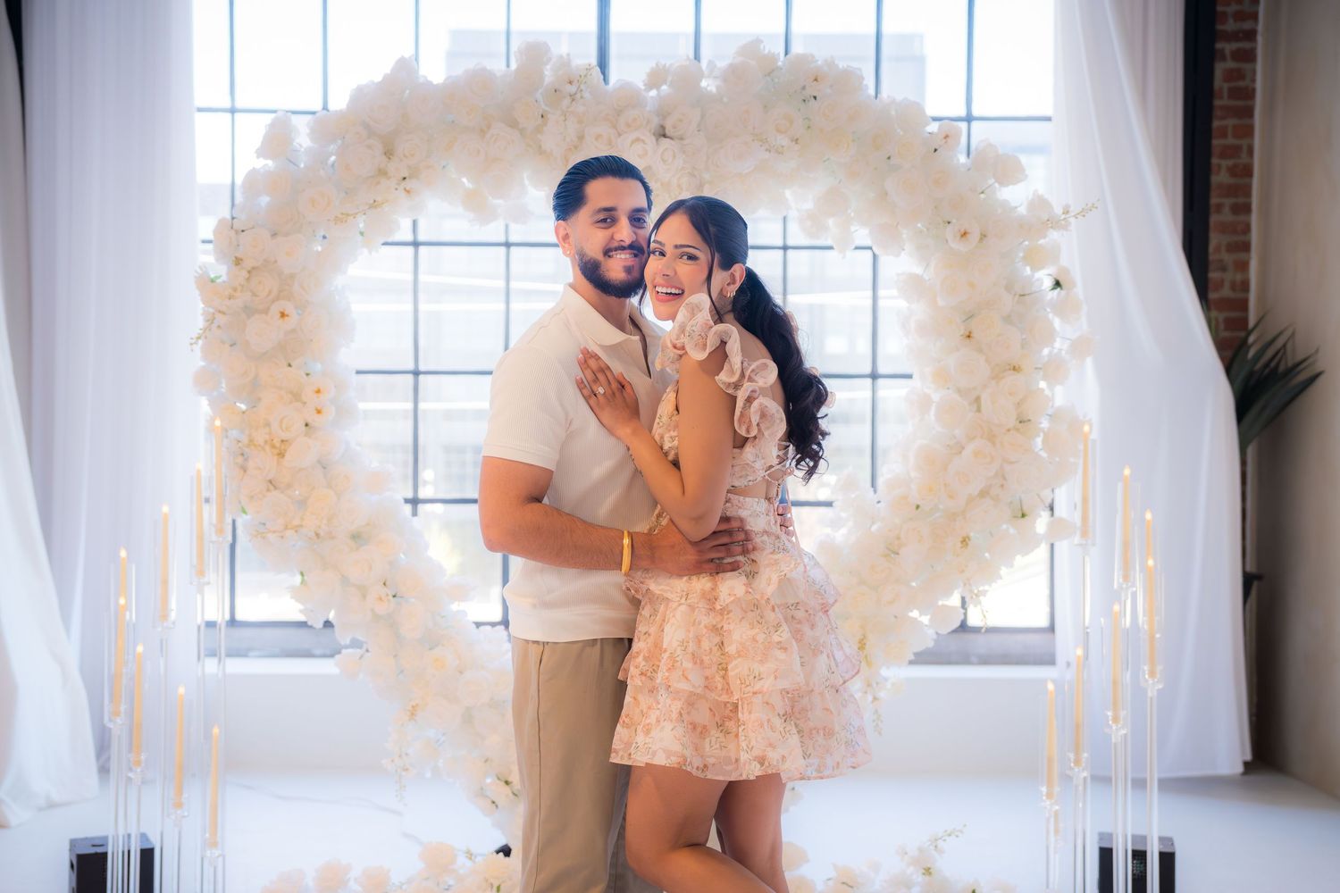 Smiling newly engaged couple posing together in front of a large white floral heart arch with tall candles.