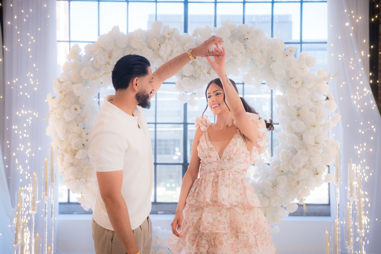 Man spinning woman in floral dress under white heart arch with twinkling lights.