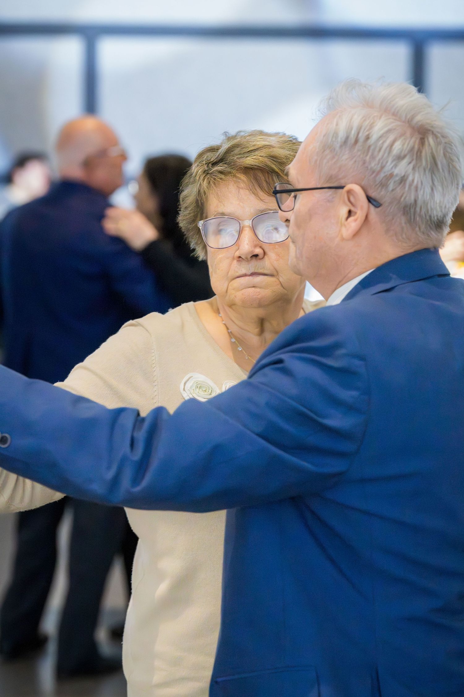 Elderly couple dancing together at a formal event, man in blue suit, woman in beige sweater wearing glasses.