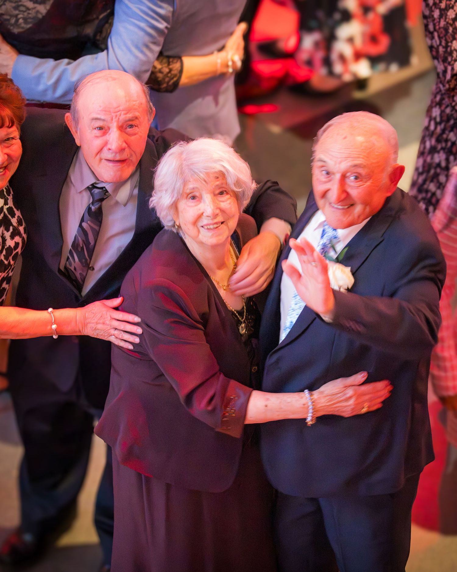 Three elderly people embracing and smiling at a festive celebration, with guests visible in the background.