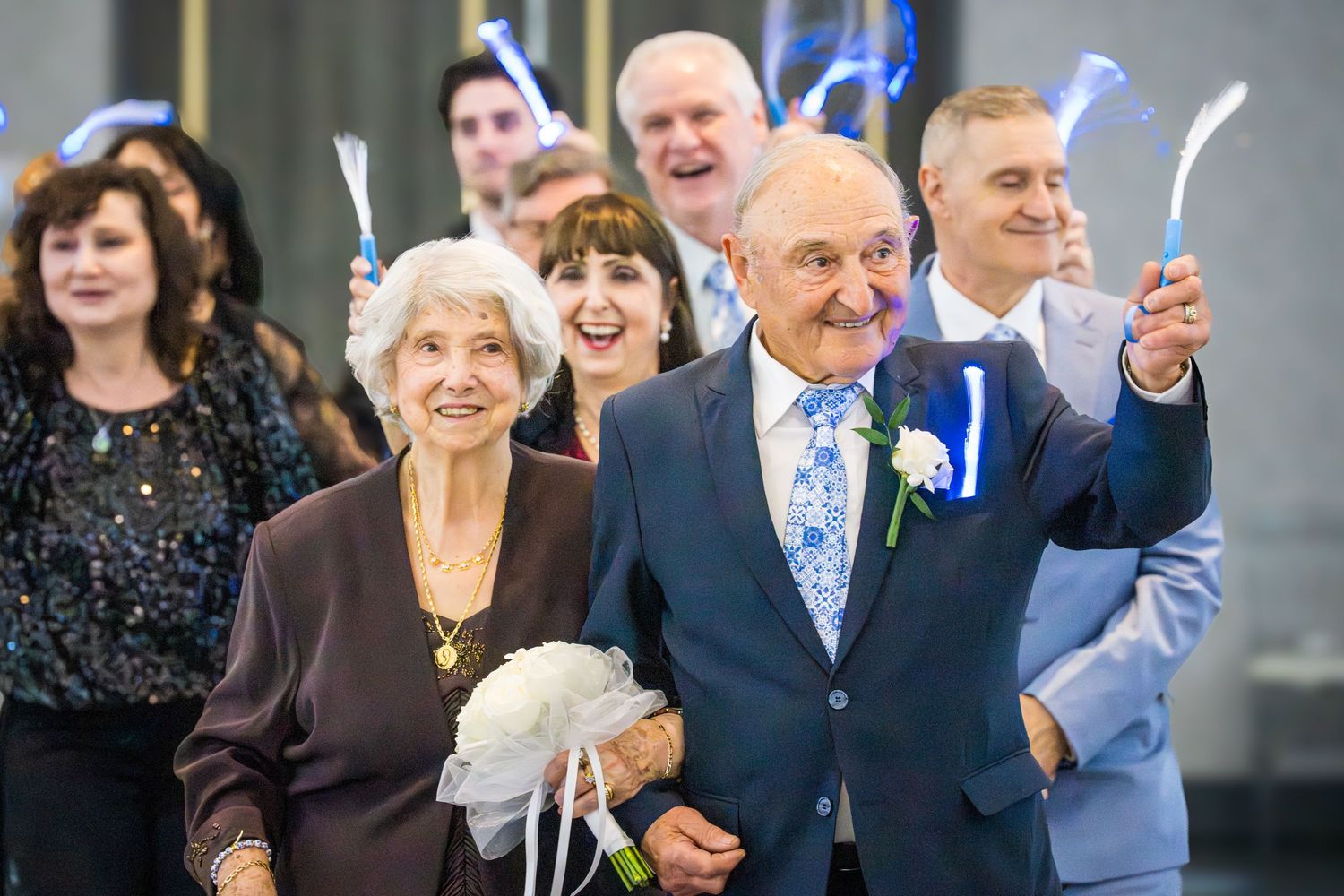 Elderly couple holding light wands leading a joyful wedding anniversary procession with smiling guests.