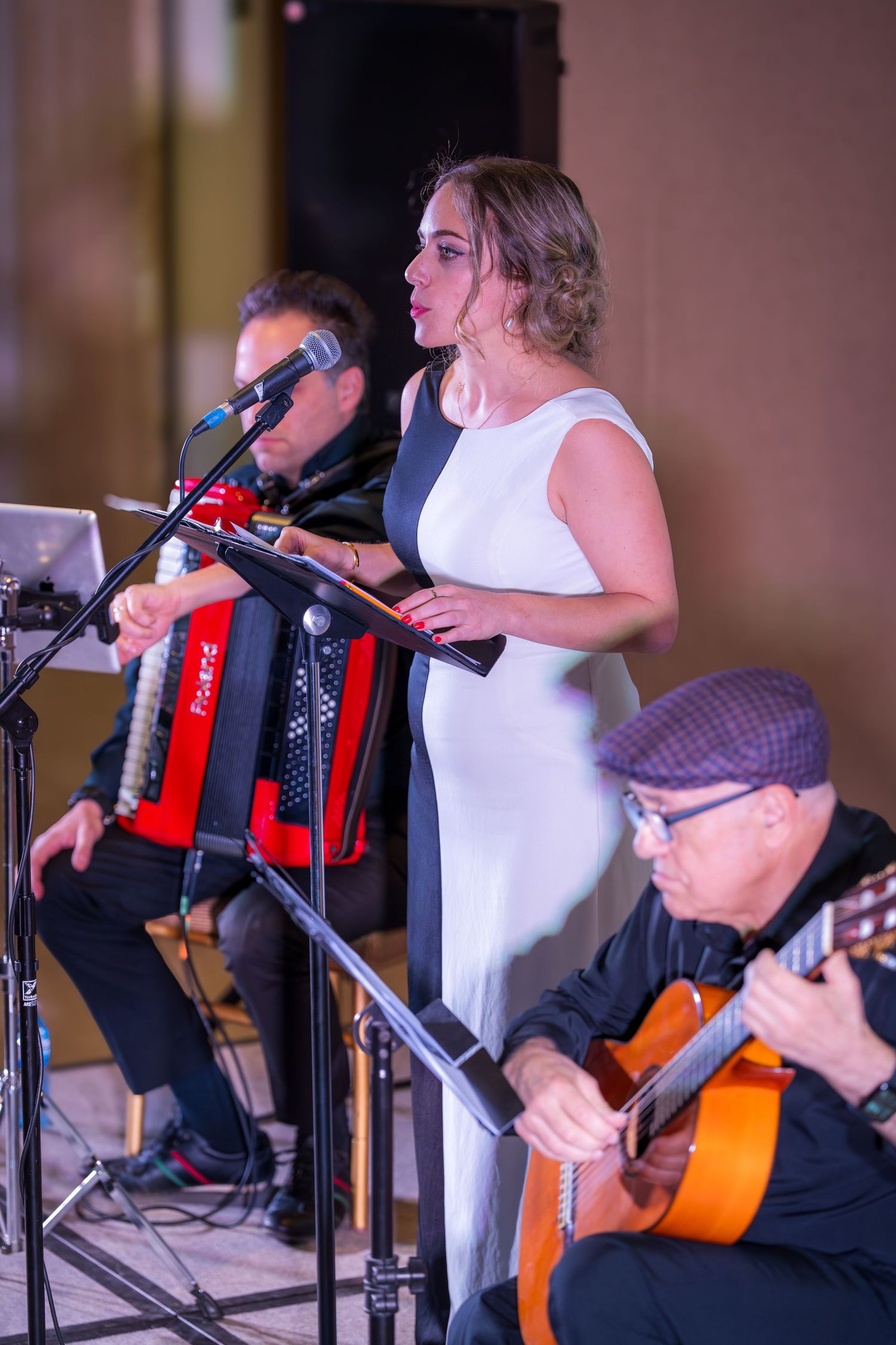 Female vocalist in black and white dress performs at microphone with accordion and guitar musicians behind her.
