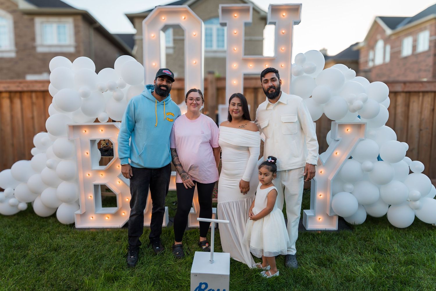 Group of five people posing at a gender reveal party with illuminated BABY marquee letters and white balloons.