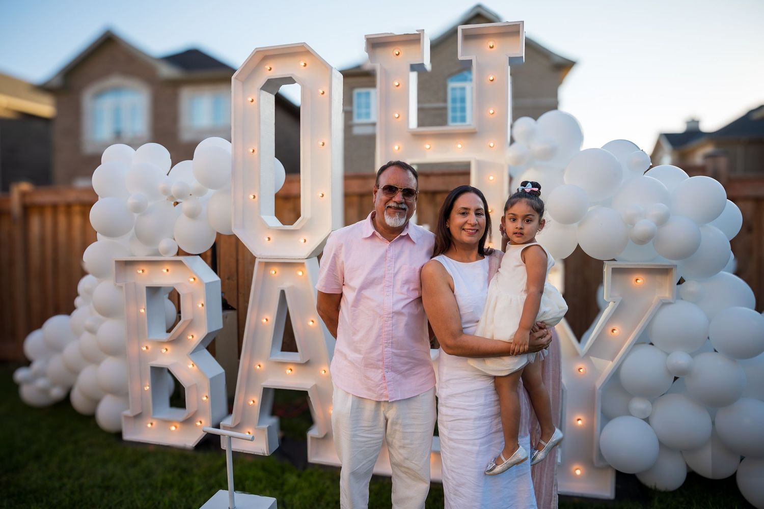 Family of three smiling in front of illuminated OH BABY marquee sign with white balloon decor.