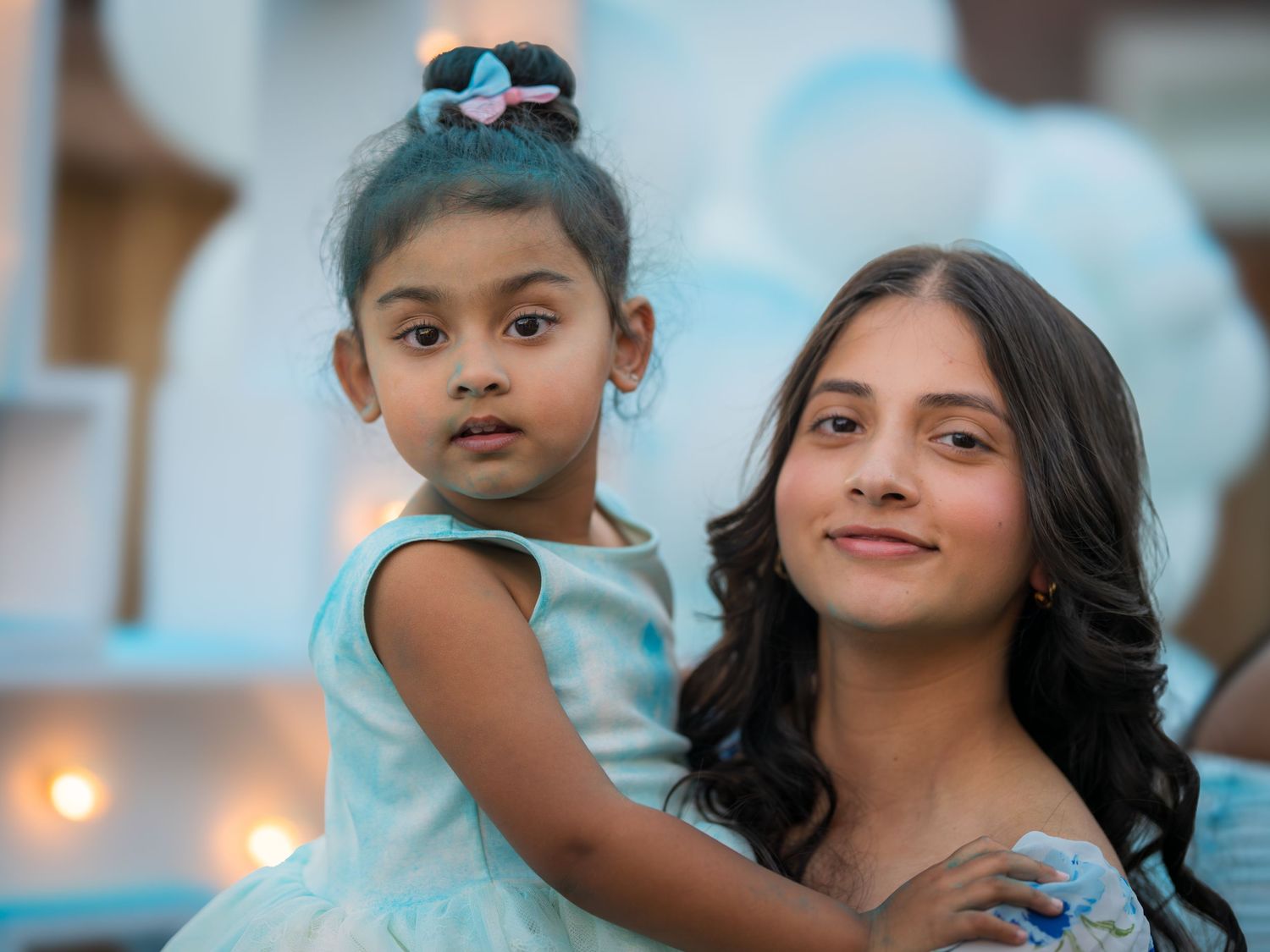 Young woman holding a little girl in blue dress in front of decorative baby shower backdrop.