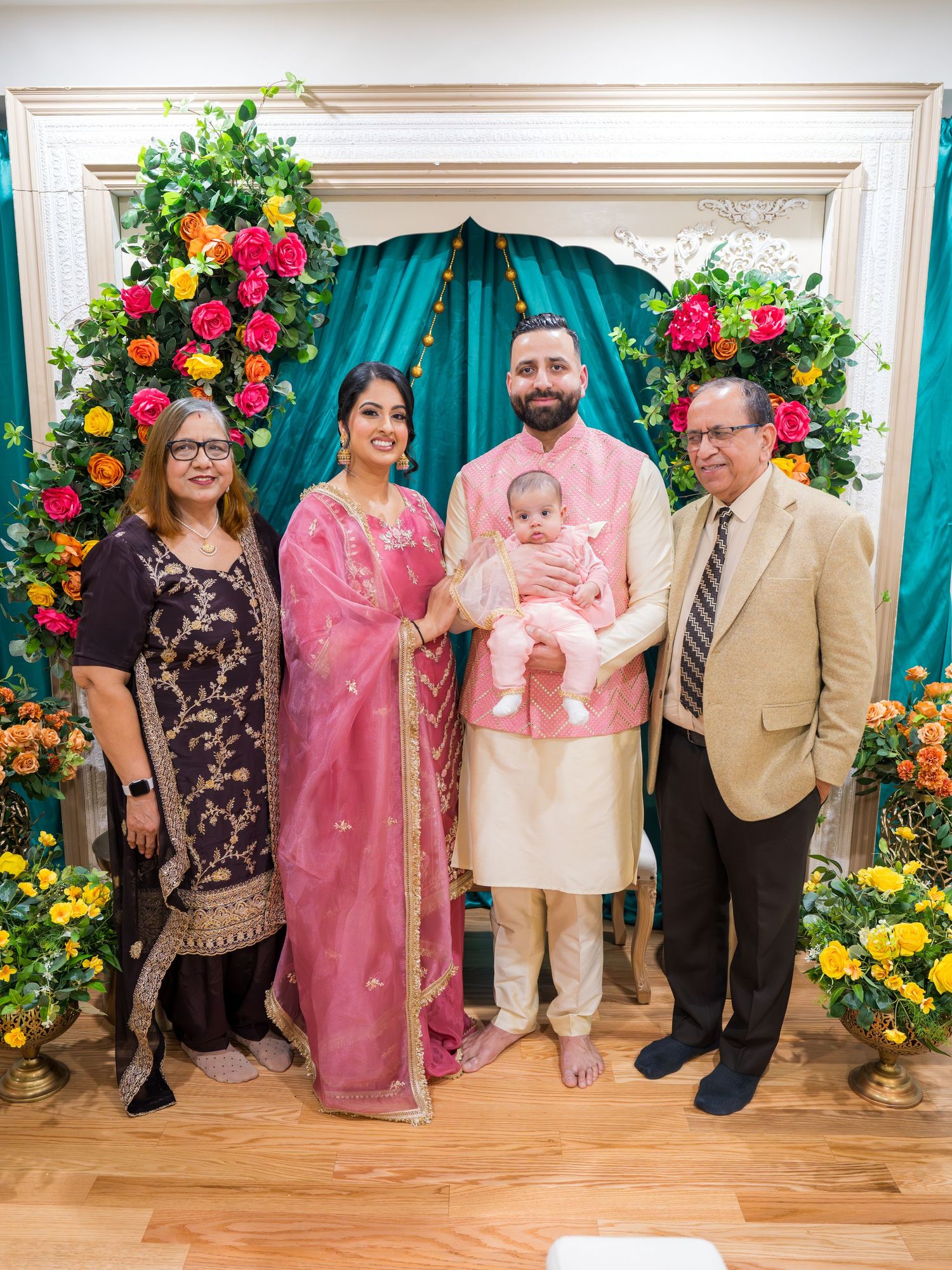 Multi-generational South Asian family with baby posing at colorful floral ceremony backdrop.