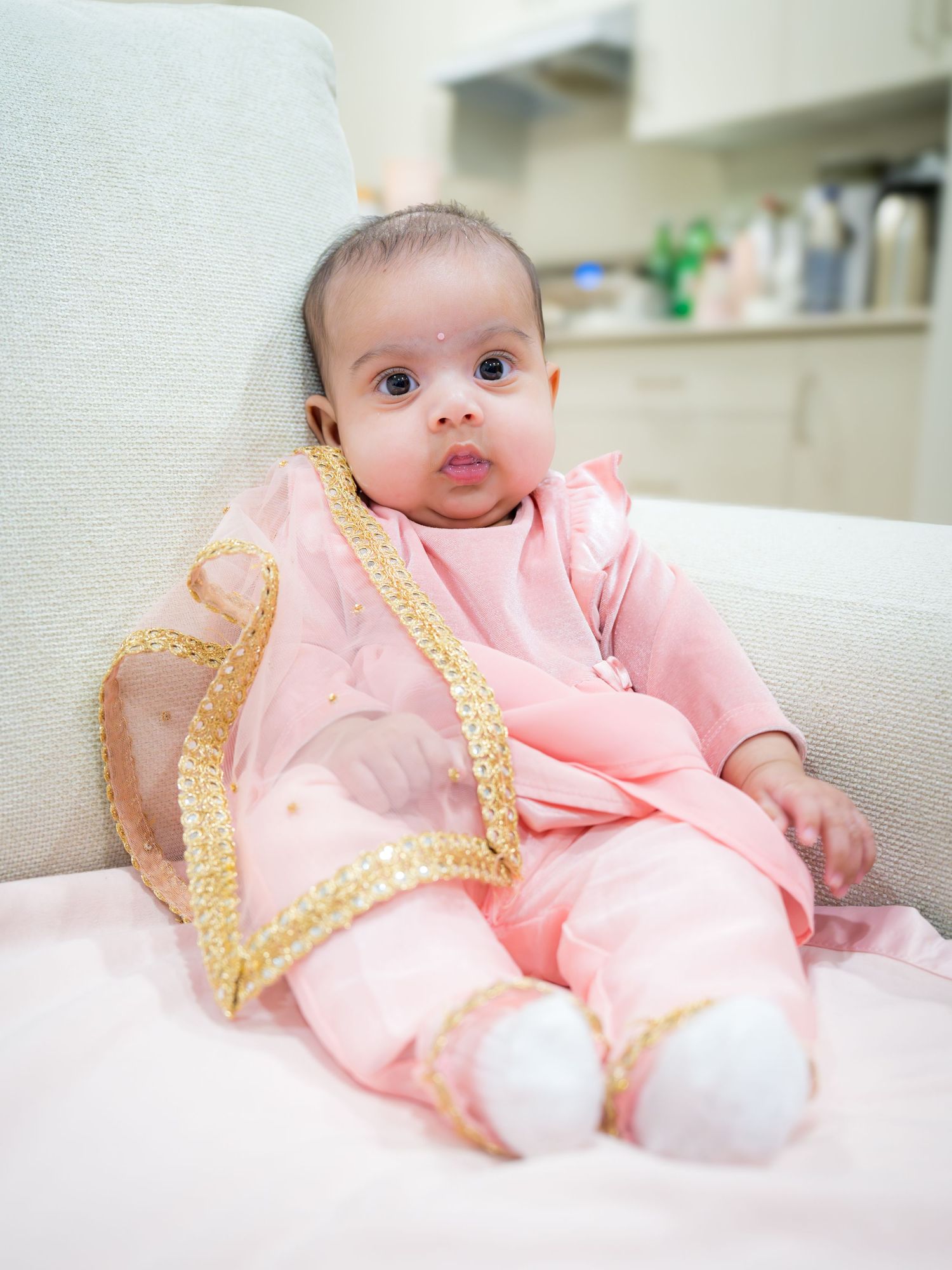Adorable baby girl in pink and gold traditional Indian outfit sitting on white sofa.
