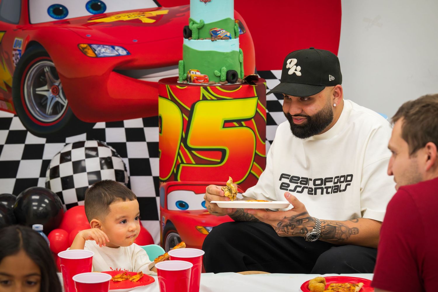 Man in white graphic tee eating with a toddler at a Cars-themed birthday party with red cups.