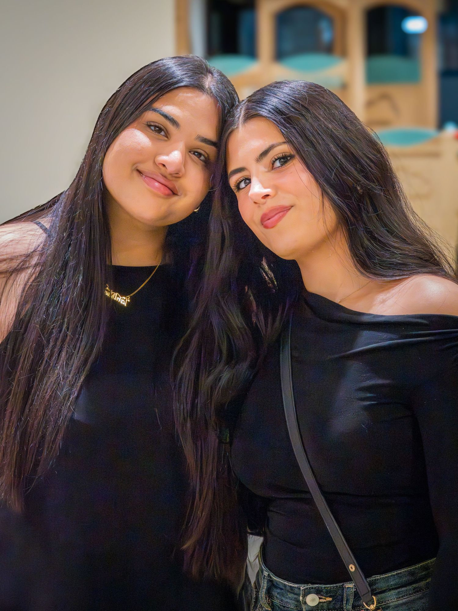 Two young women with long dark hair wearing black outfits smiling together at an indoor event.
