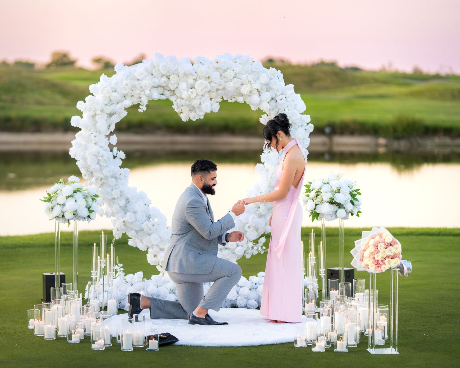 Romantic outdoor proposal setup with white floral arch and candles on a lush green golf course at sunset.