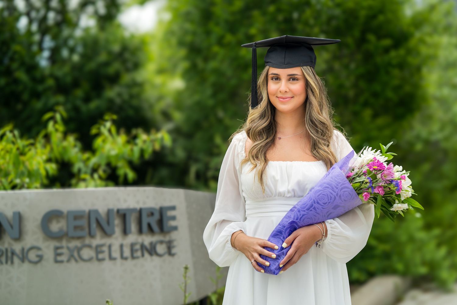 Graduate poses with flowers outside education center in white dress and cap.