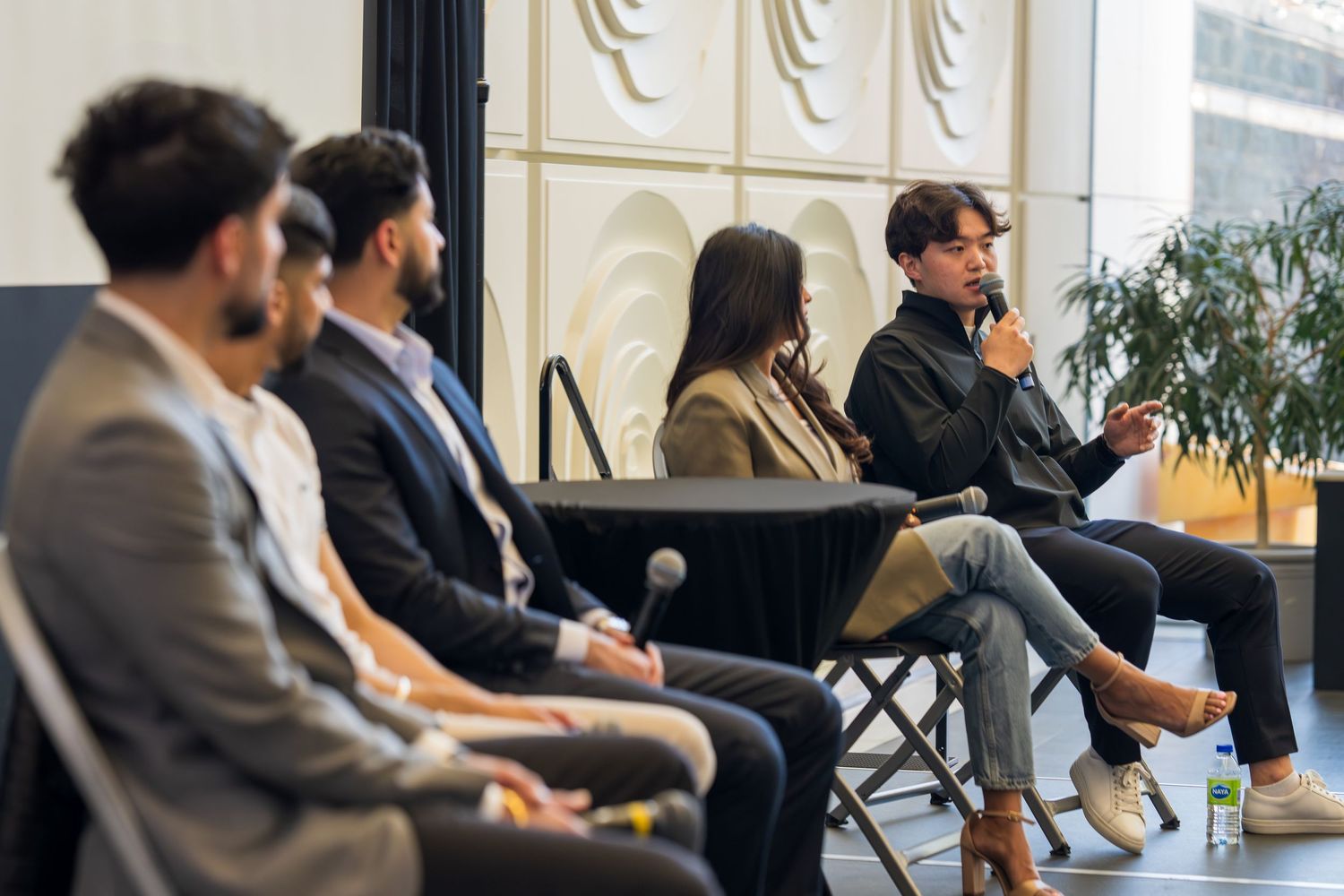 Conference panel members seated in discussion against white textured wall backdrop.