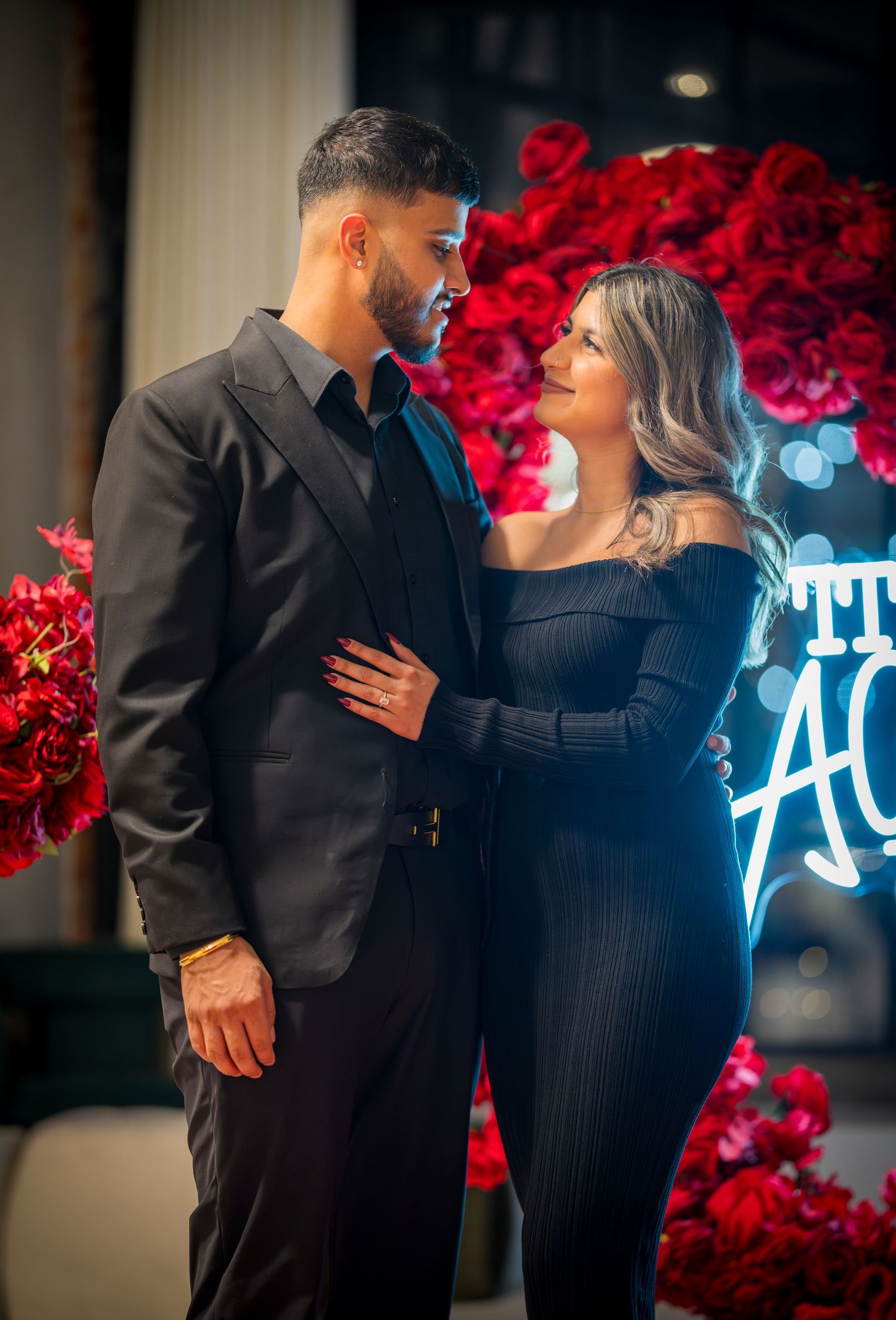 Couple in black attire share a romantic moment against a backdrop of roses and neon signage.