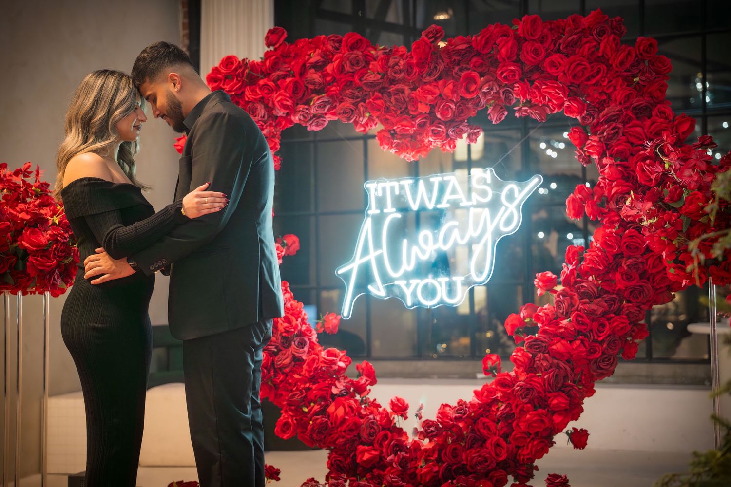 Elegant proposal setting with red rose wall, neon lights, and scattered rose petals.
