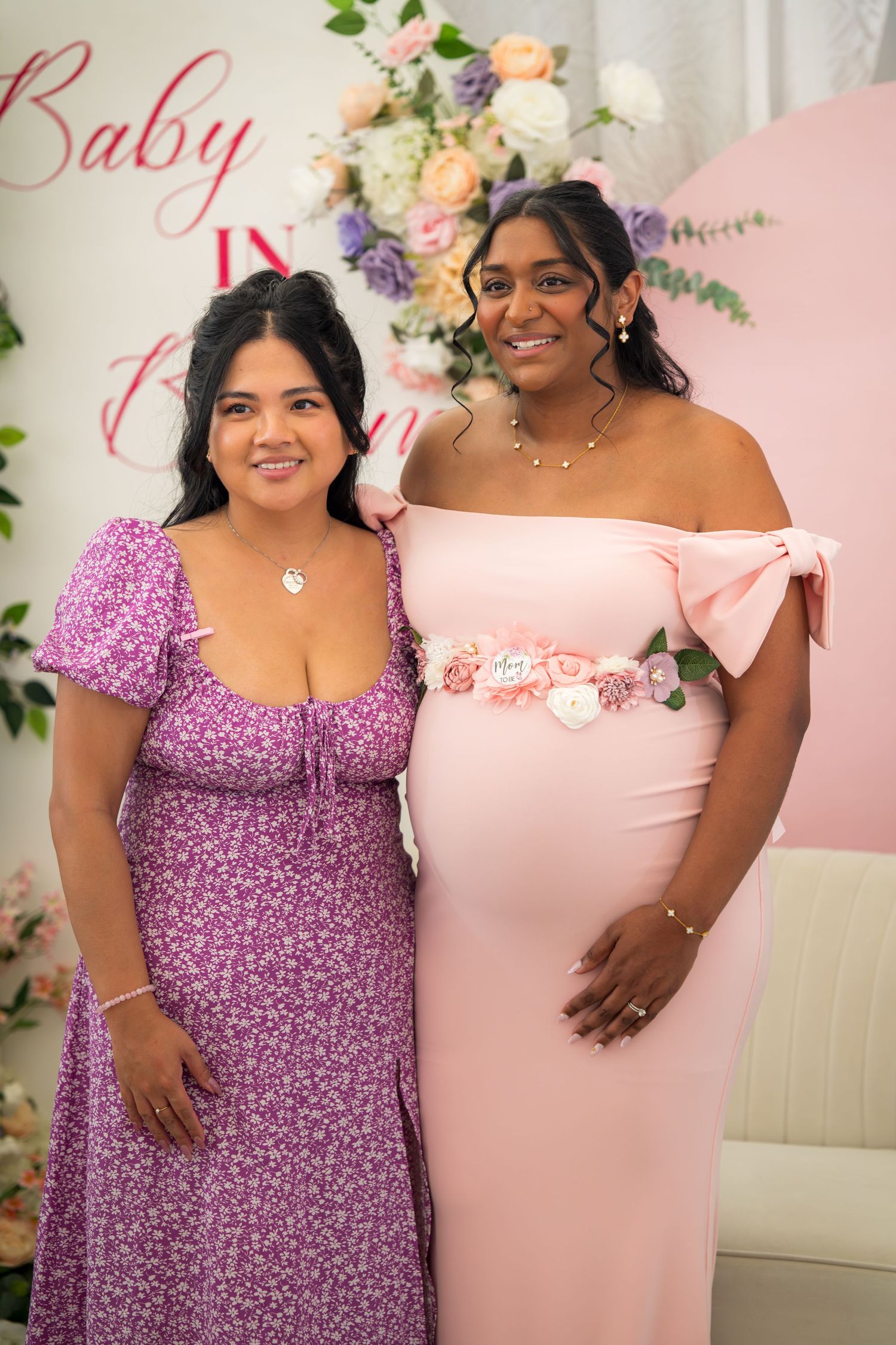 Two guests pose together at a baby shower, one in purple floral dress and one in pink.