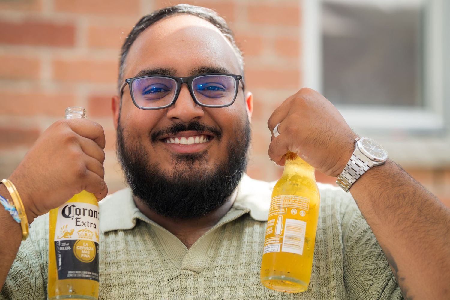 Person in glasses and polo shirt holding Corona beer bottles while smiling.