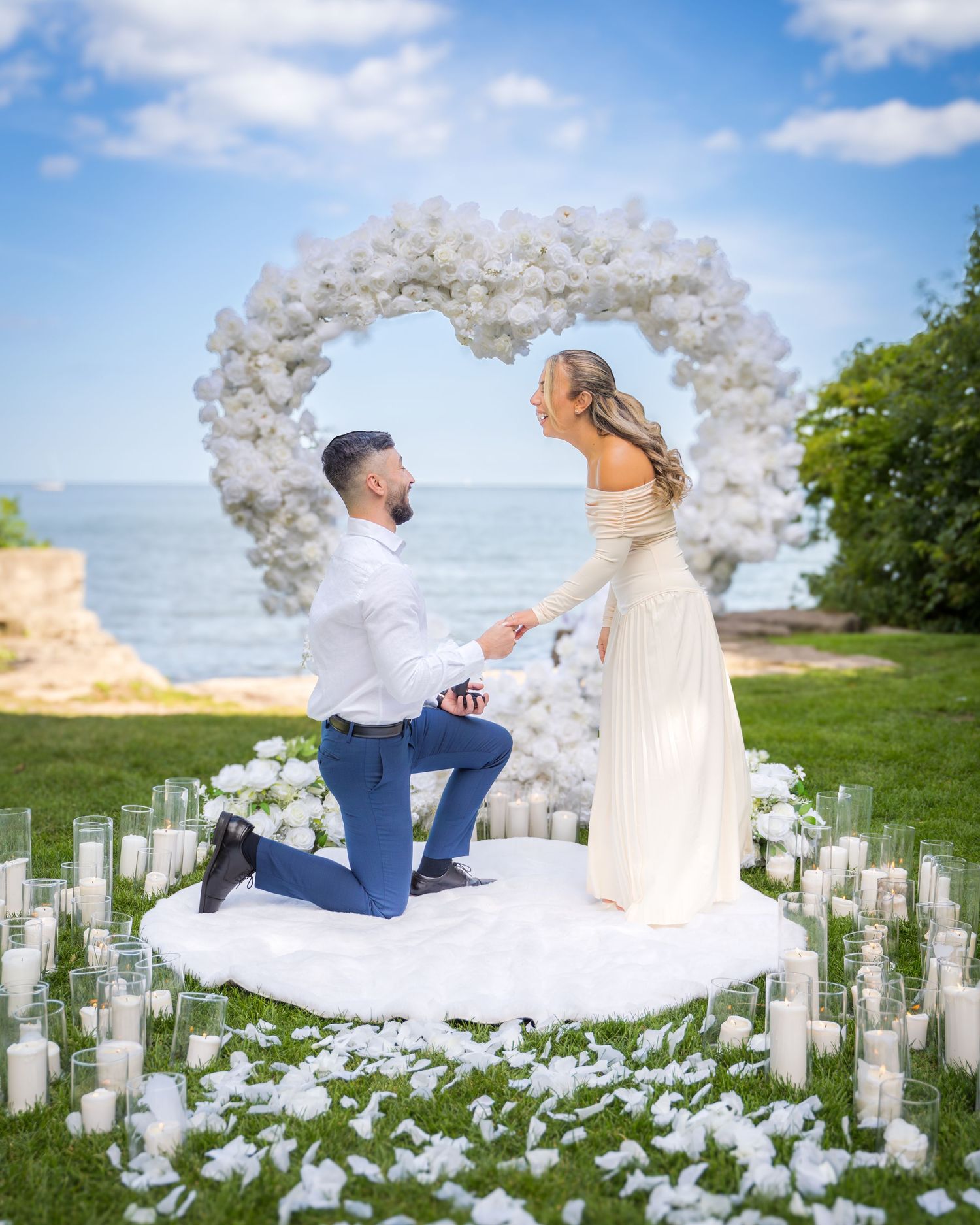 Romantic proposal setup featuring white floral arch and glass-enclosed candles on grass.