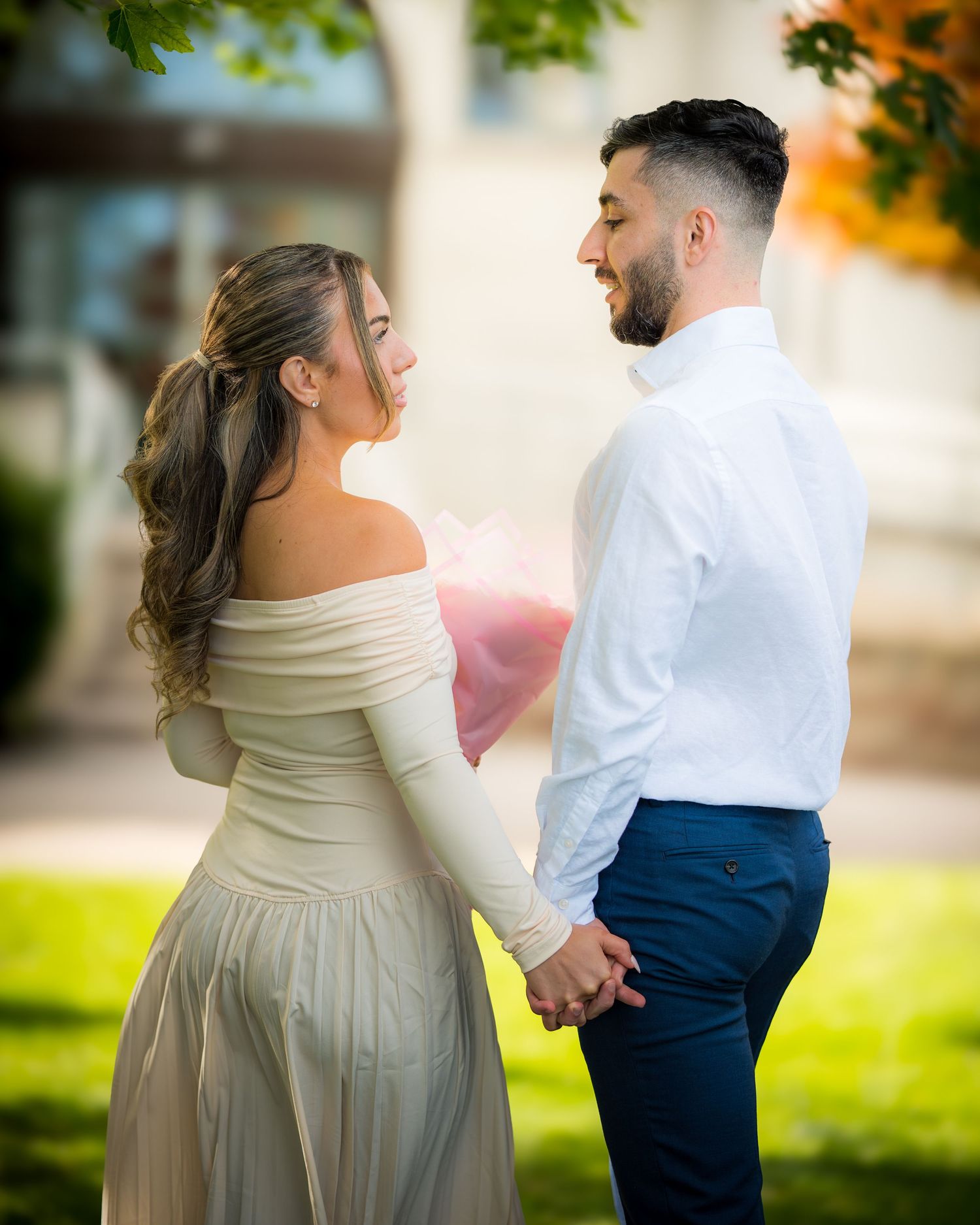 Couple in formal attire share a romantic moment outdoors during a wedding photoshoot with autumn foliage in background.