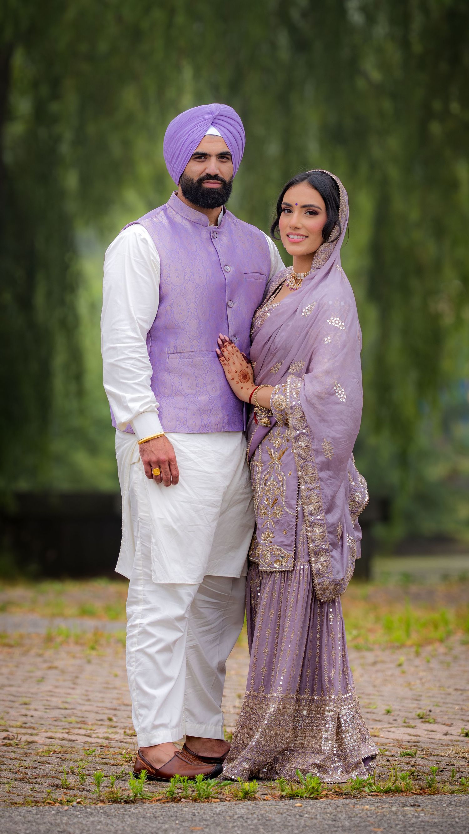A couple in coordinated lavender and white traditional Punjabi outfits poses together on a leaf-covered path.