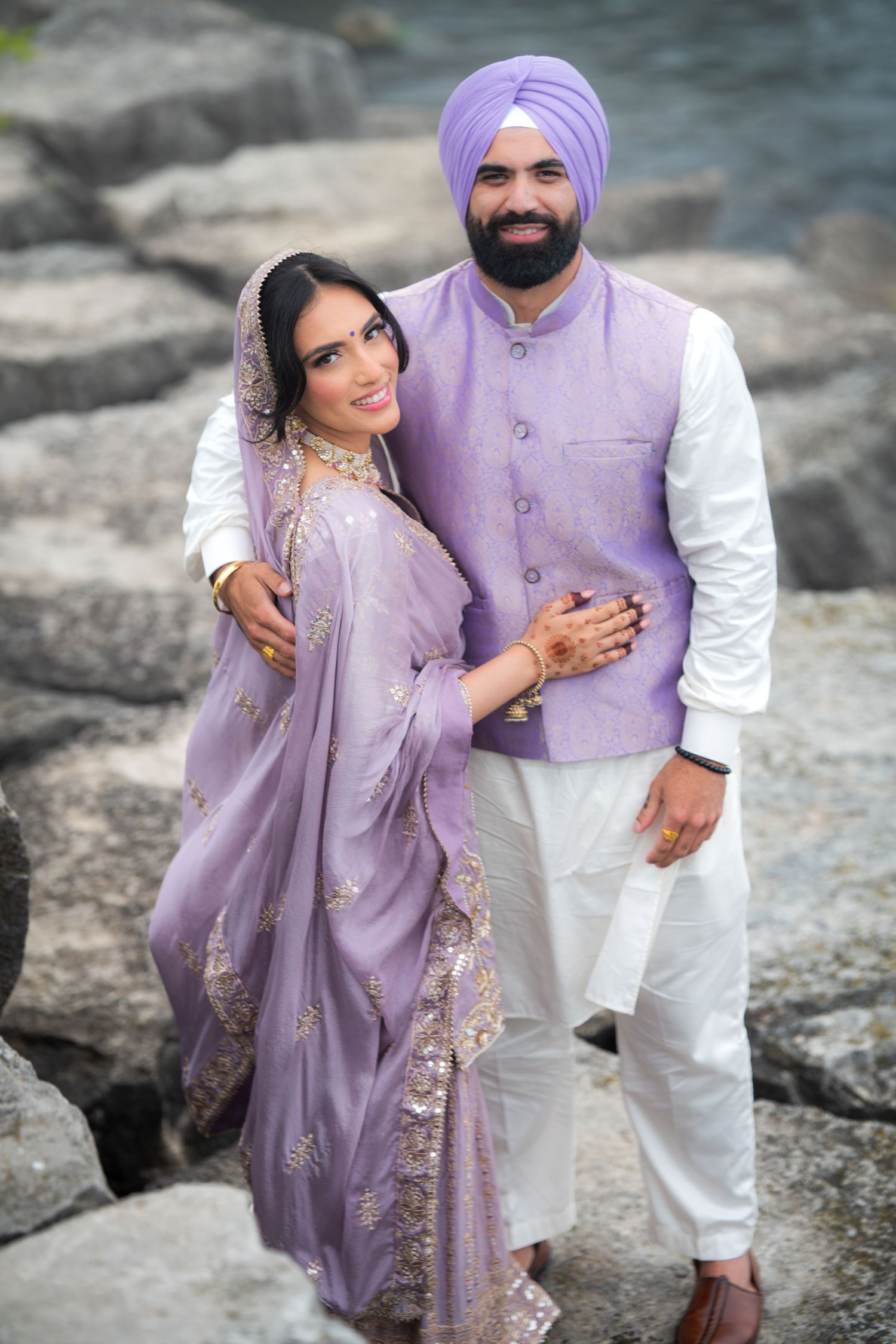 Couple wearing coordinated purple and white traditional outfits pose on rocky terrain.