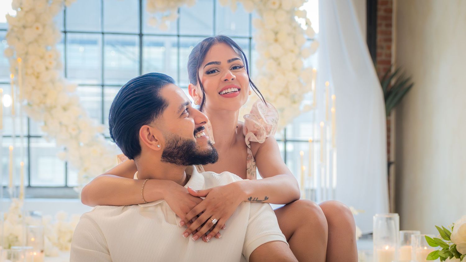 Engaged couple sitting closely together smiling in front of a white floral heart-shaped arch.