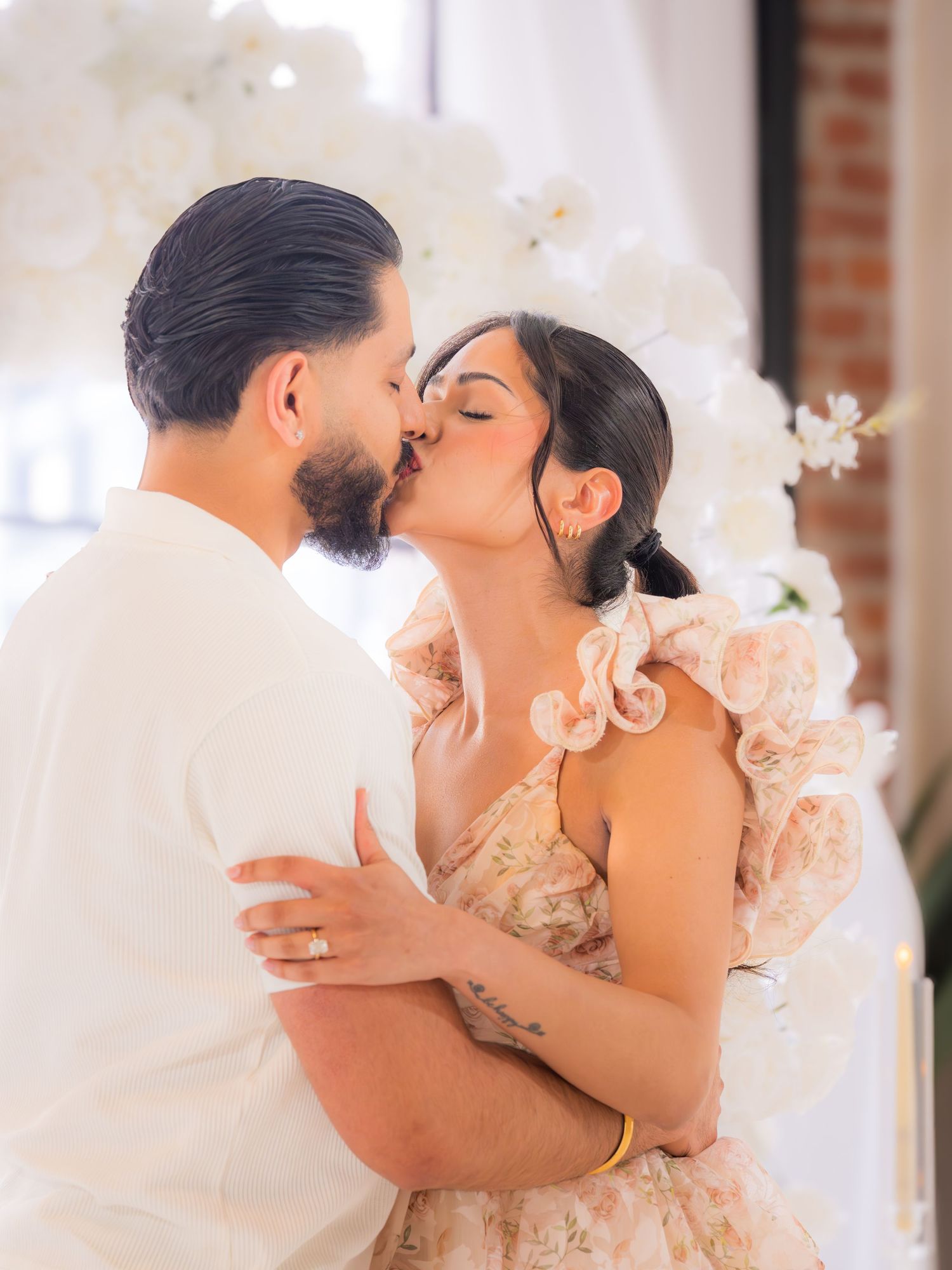 Couple sharing a romantic kiss in front of white orchid floral backdrop at elegant engagement celebration.