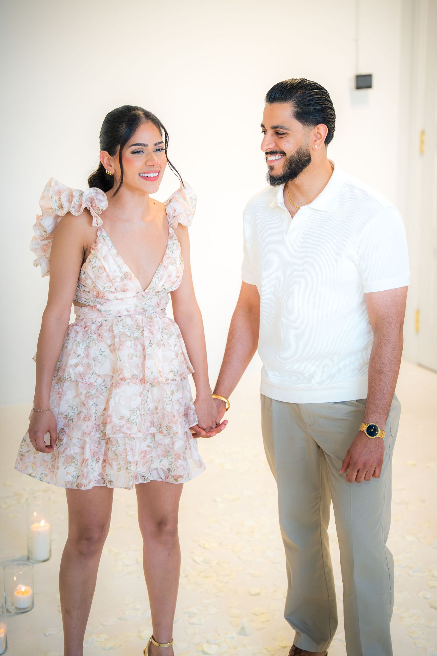 A smiling couple holds hands and gazes at each other surrounded by candles in a bright white room.