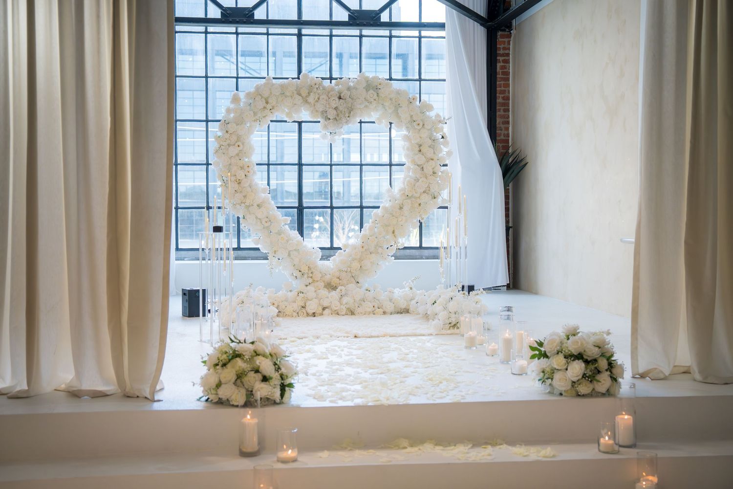 Elegant white floral heart arch wedding proposal setup with candles and rose petals.