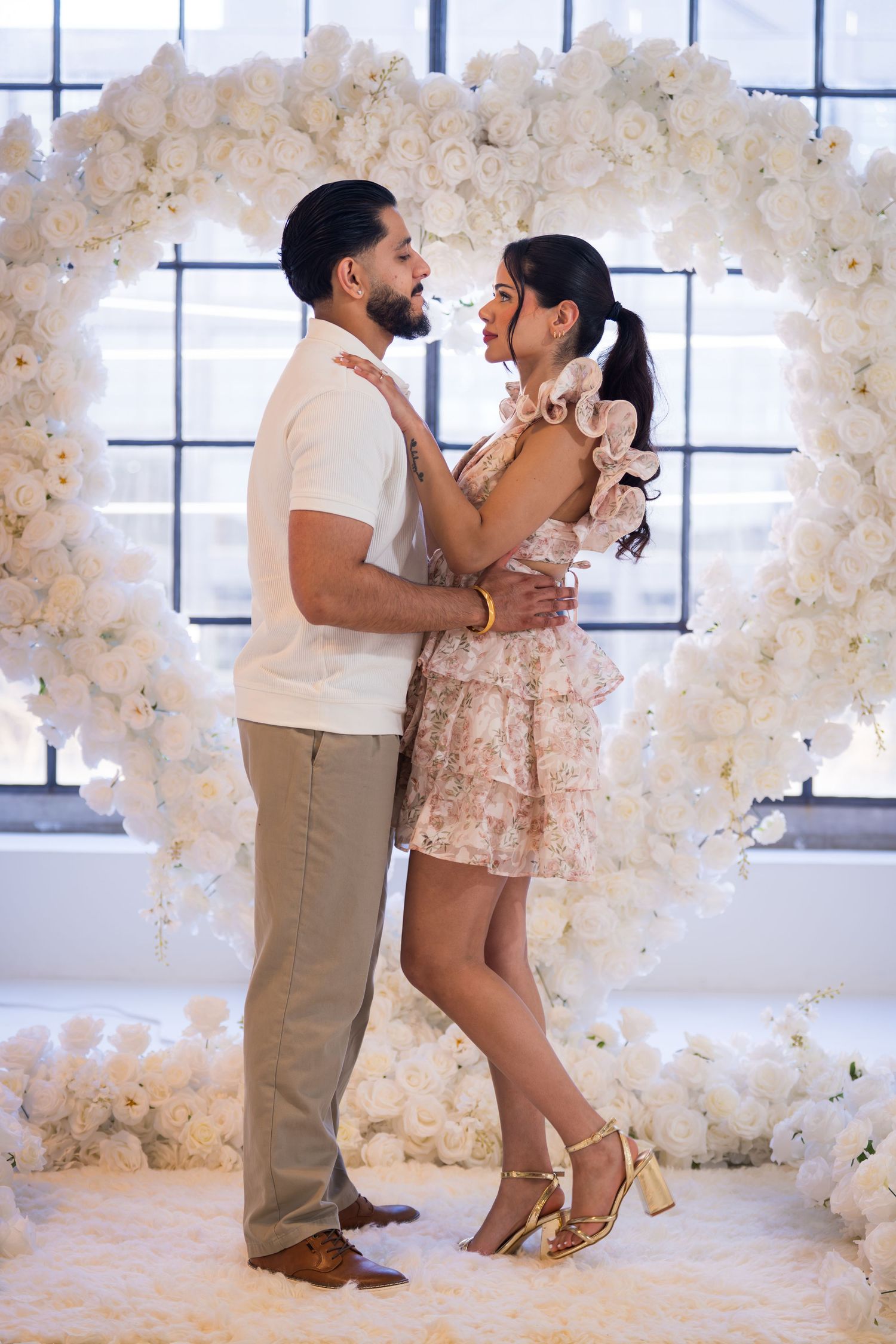 Couple embracing lovingly inside a large heart-shaped white rose arch near industrial windows.
