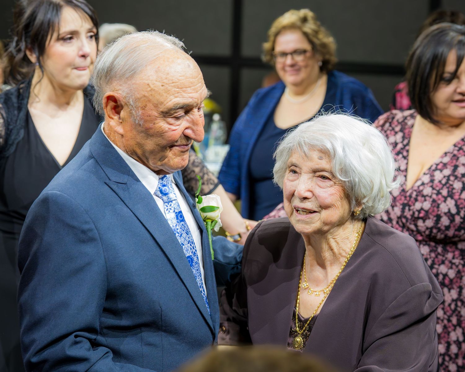 Elderly man in blue suit with boutonniere chatting warmly with an older woman at a celebratory gathering.