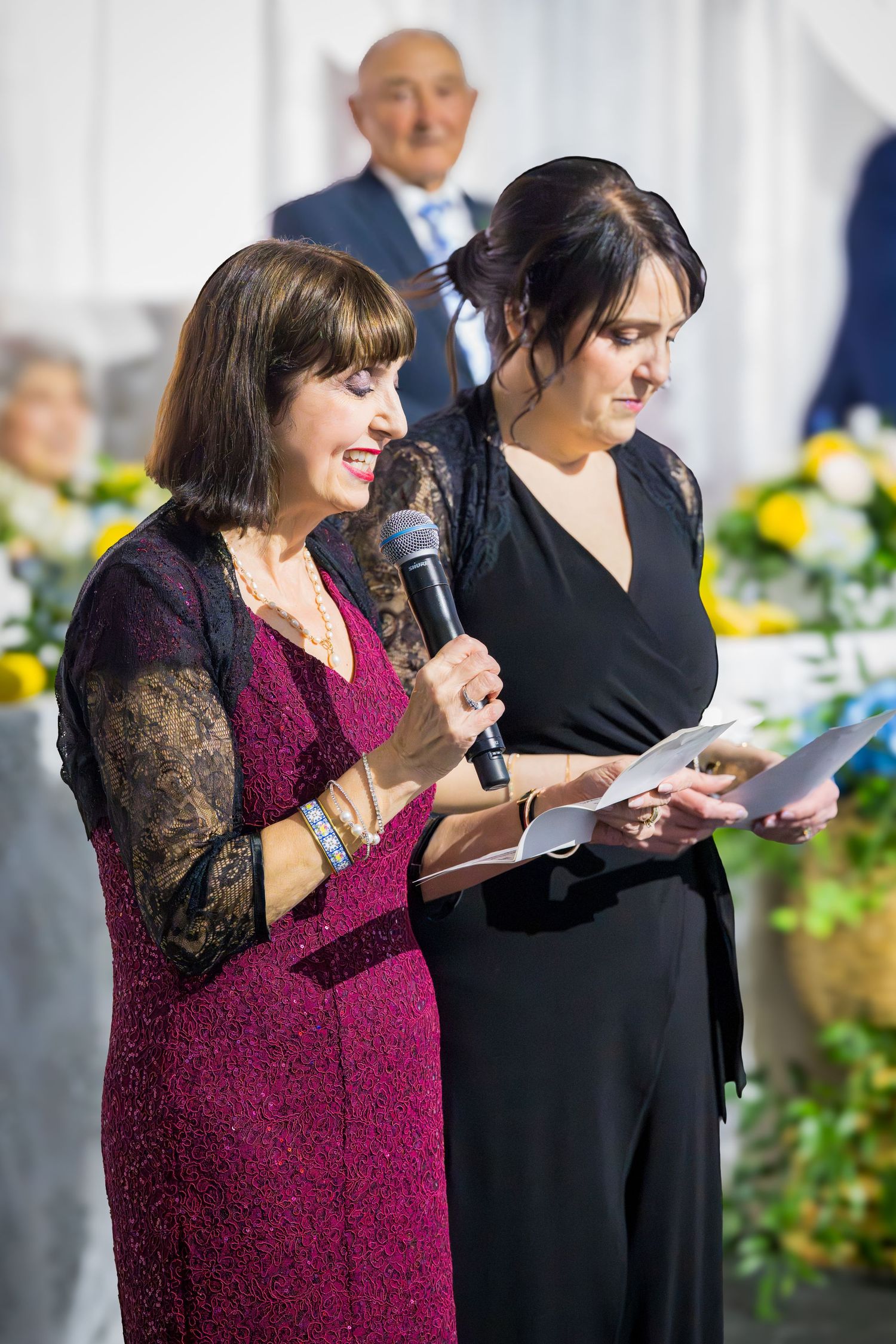 Two women at a formal event, one speaking into a microphone in a purple dress while the other holds papers in black.