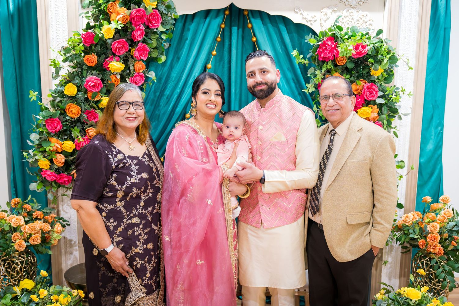 Family of four posing with baby at colorful floral backdrop during Indian Lohri celebration ceremony.