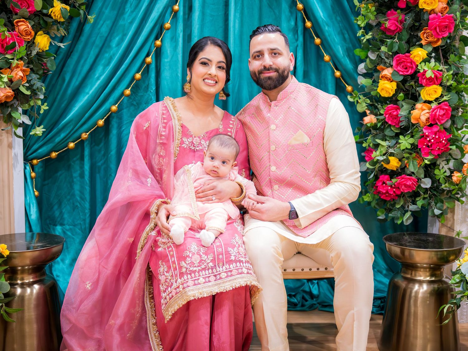 Indian couple in coordinating pink traditional wear seated with their baby at a decorated cultural ceremony.