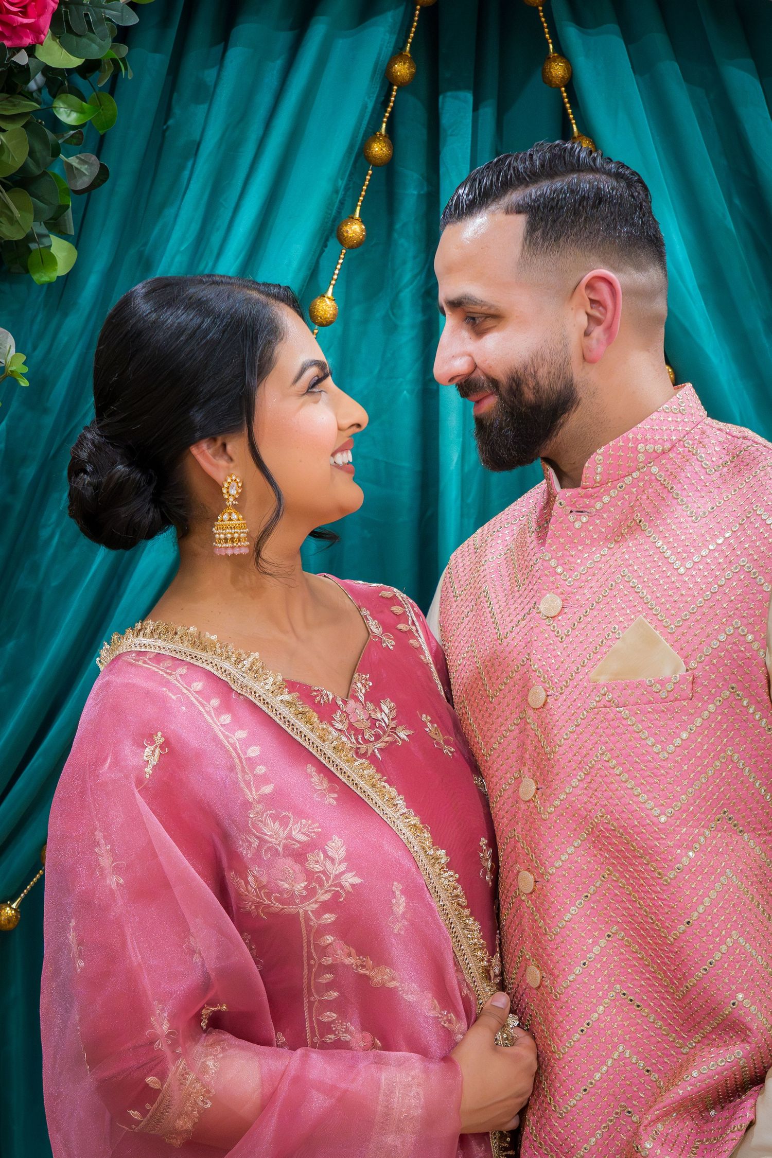 A couple in matching pink Indian traditional outfits smiles at each other against a teal draped backdrop.
