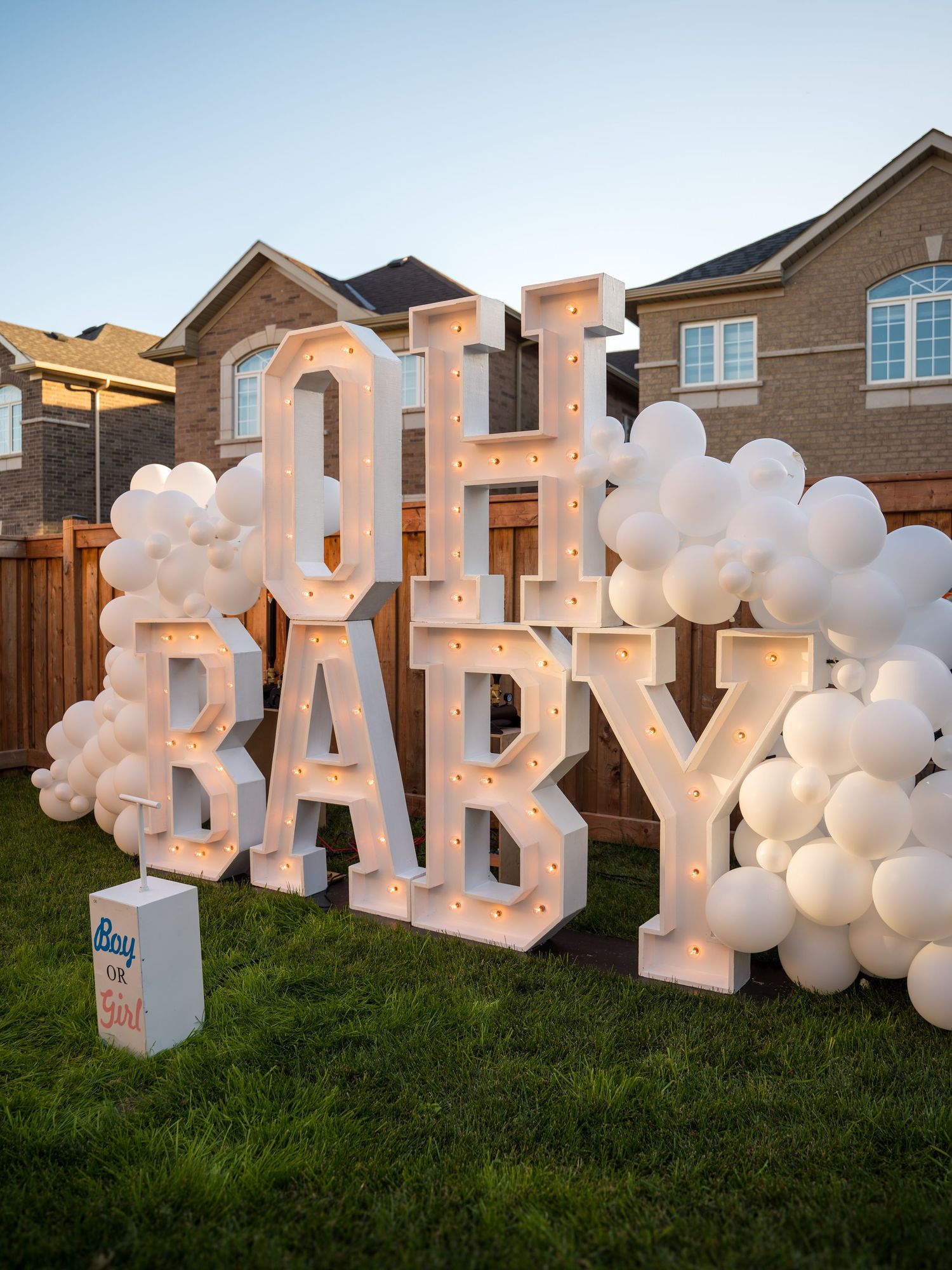 Illuminated OH BABY marquee letters with white balloon garland on grass for an outdoor baby shower gender reveal.