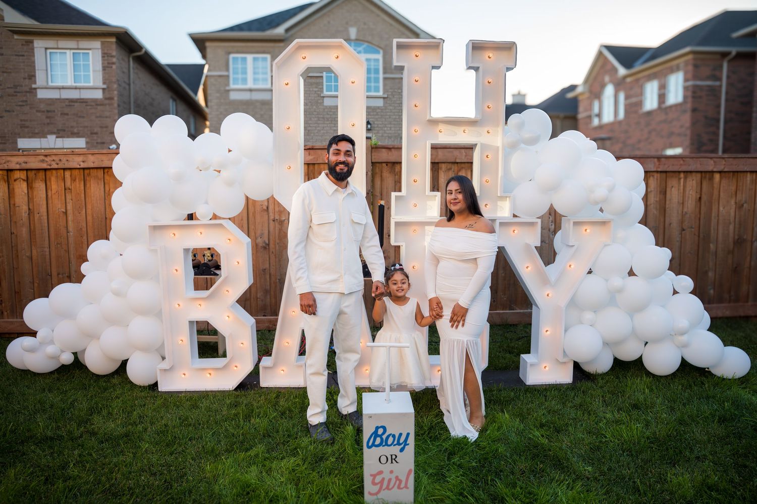 A couple and child stand before illuminated OH BABY marquee letters at a backyard gender reveal.