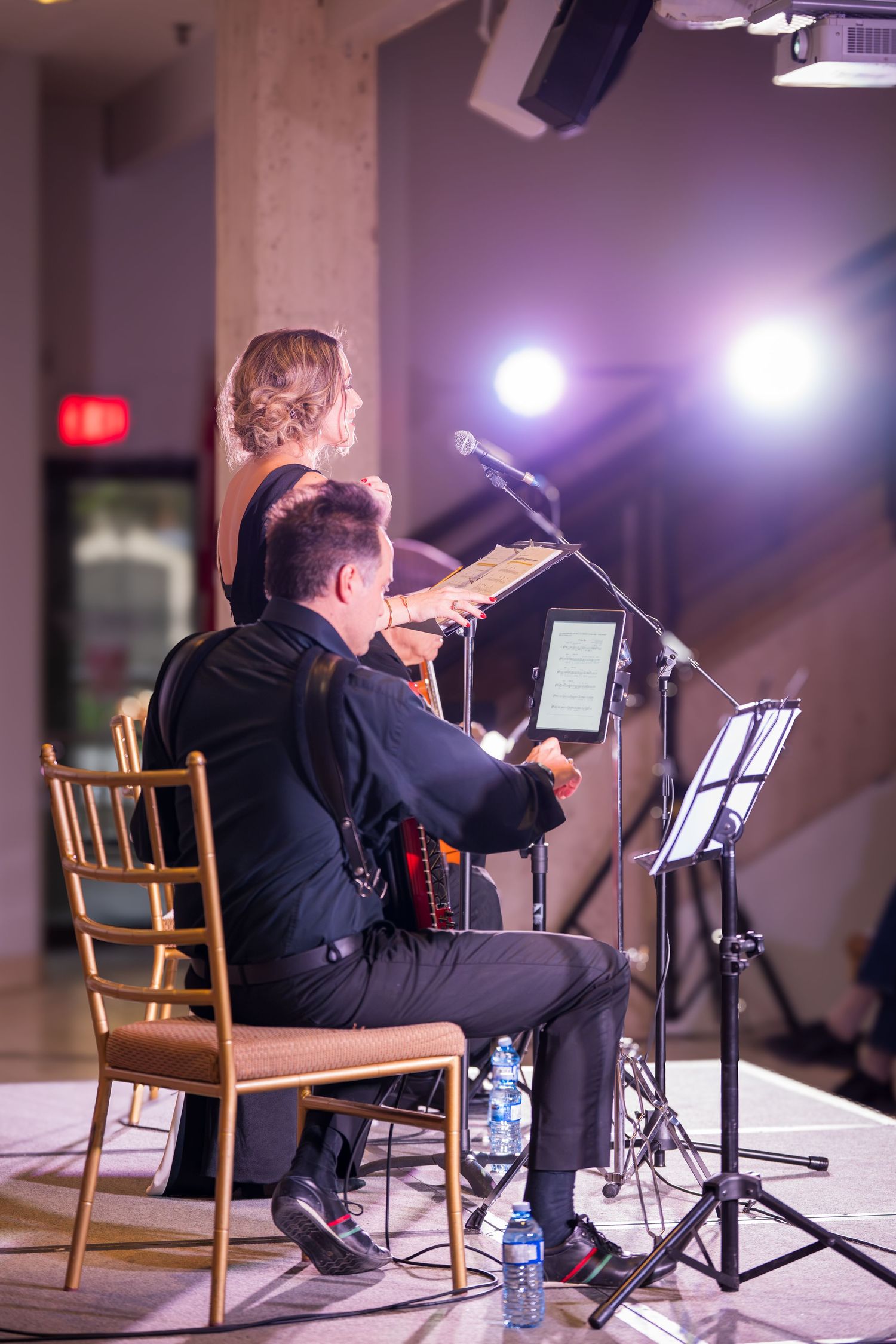 A musician plays violin while seated on stage next to a standing woman reading from a music stand under bright lights.