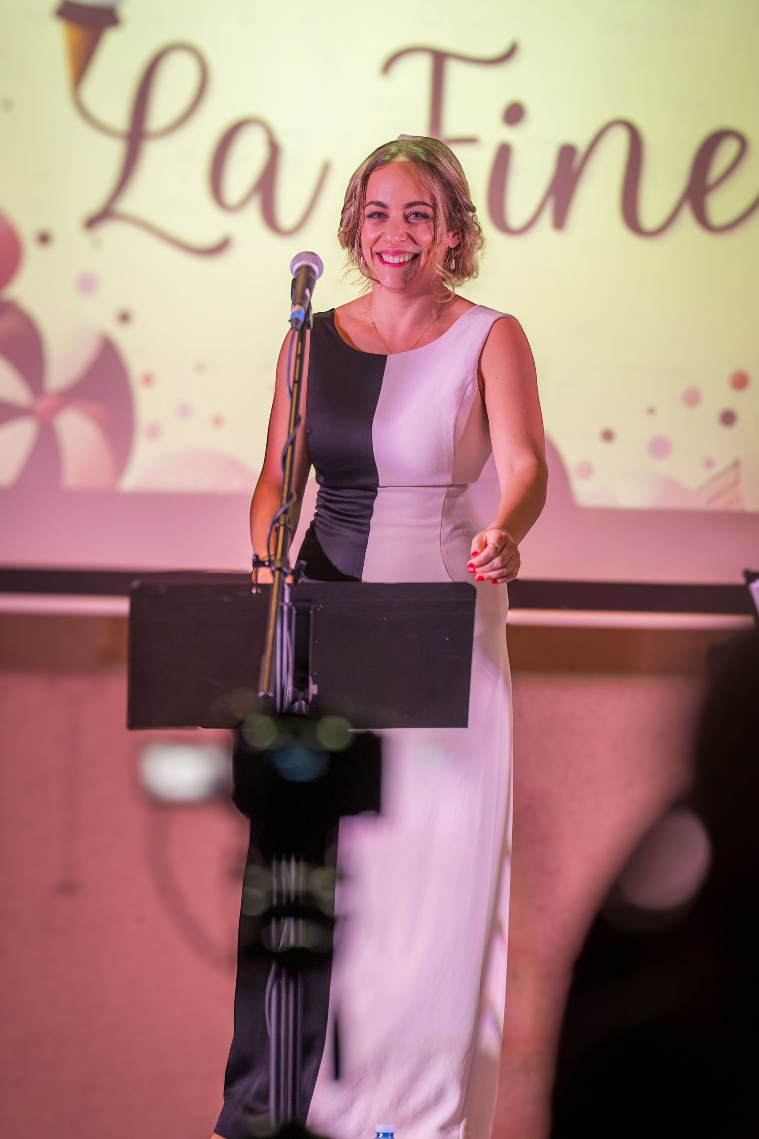 Smiling woman in black and white gown speaks at podium during La Fine themed event.