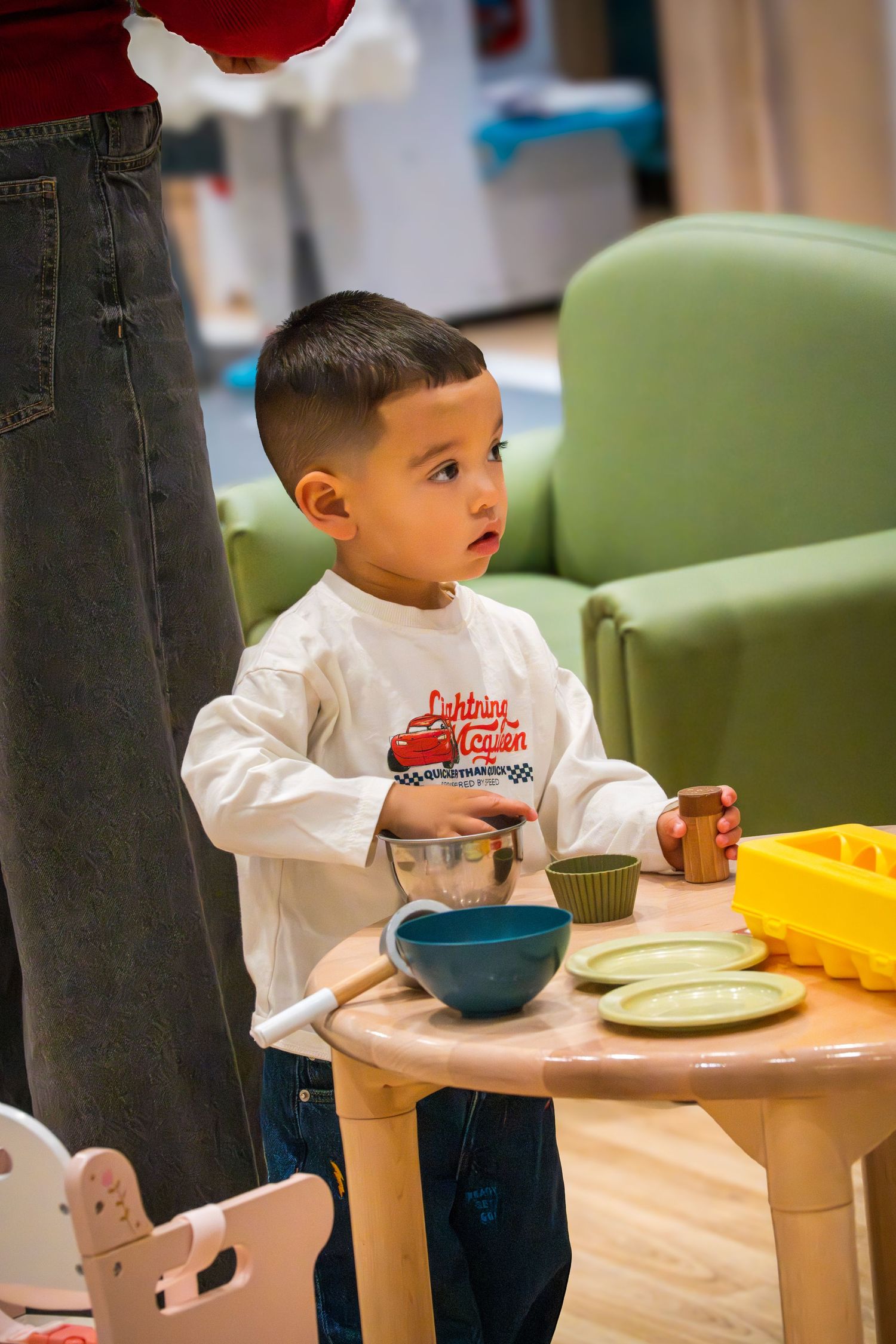 Young boy in Lightning McQueen shirt playing with toy kitchen set at small wooden table.