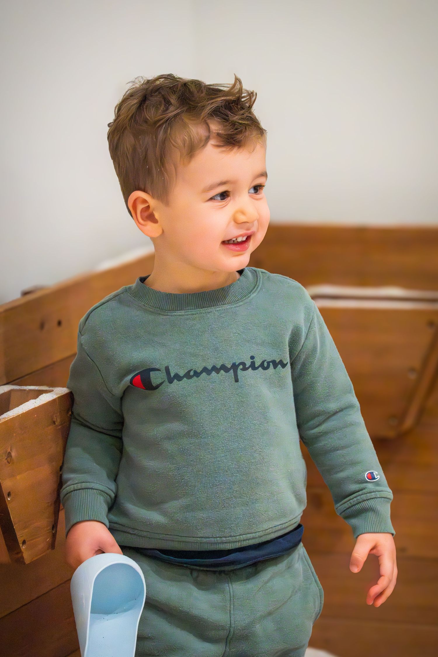 Toddler boy smiling and wearing a green Champion sweatsuit while holding a toy indoors.
